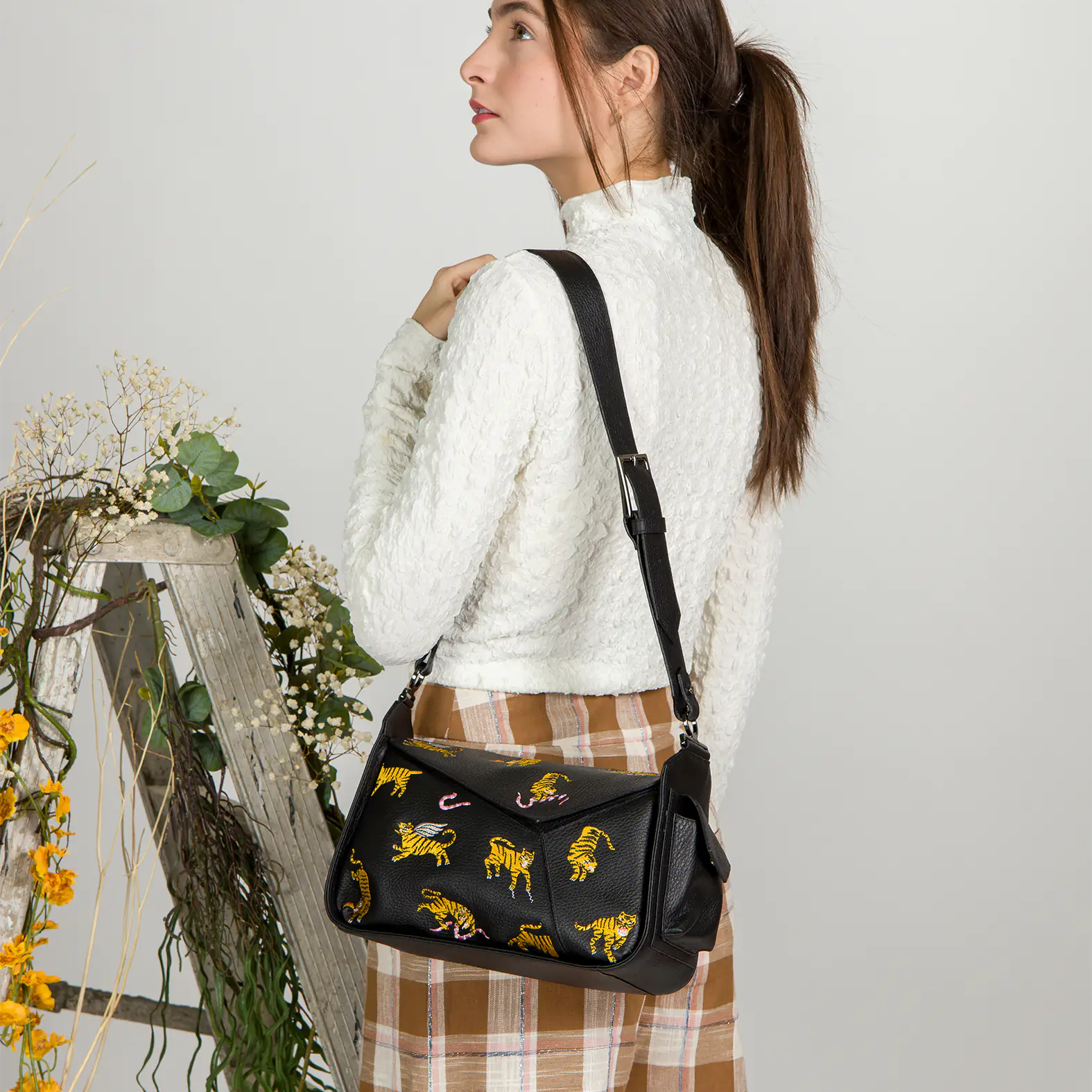 Woman in a white textured top and plaid skirt poses with a black shoulder bag featuring embroidered yellow tigers and pink snakes, styled with a wide adjustable strap.
