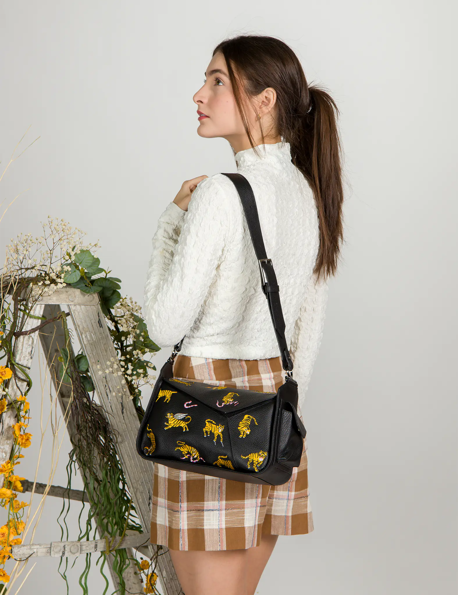 Woman in a white textured top and plaid skirt poses with a black shoulder bag featuring embroidered yellow tigers and pink snakes, styled with a wide adjustable strap.