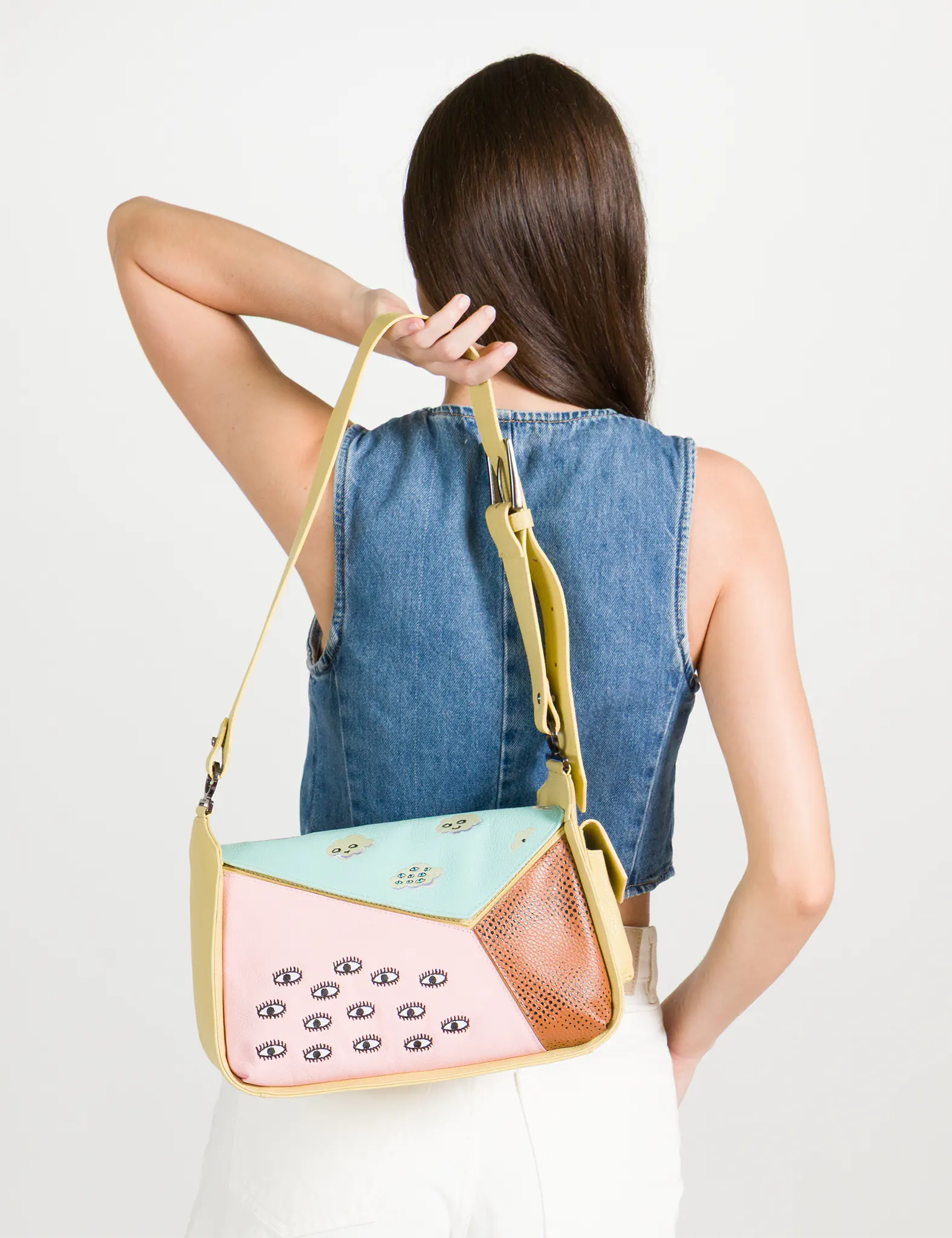 Back view of a model in a sleeveless denim top and white shorts holding a pastel color-block shoulder bag with embroidered eye motifs and an adjustable yellow strap.