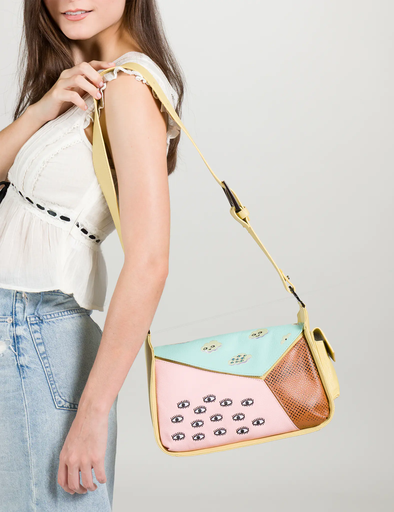 Close-up of a model wearing a white blouse and denim skirt, holding a pastel color-block shoulder bag with embroidered eyes, a yellow strap, and brown textured leather detail.