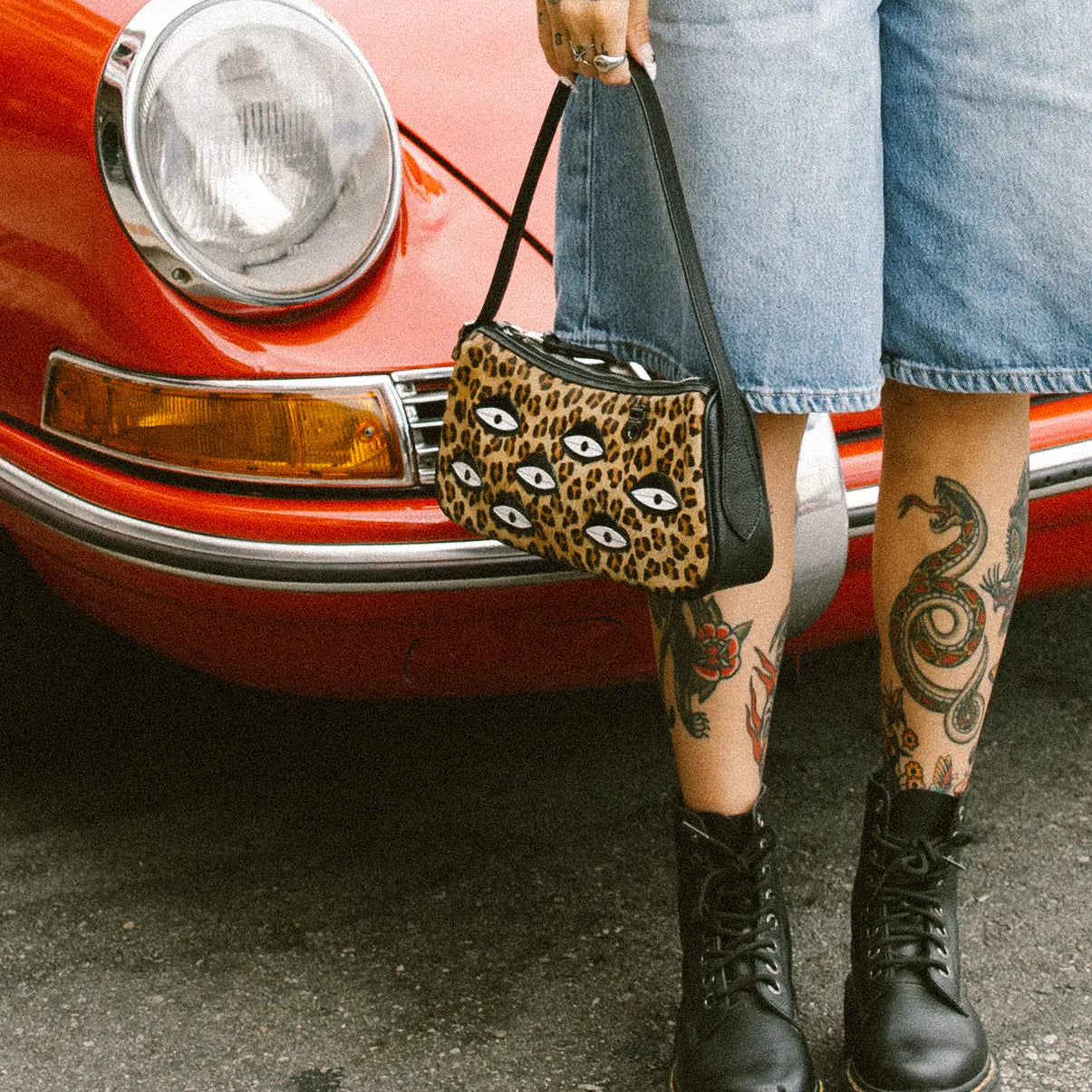 Tattooed woman in denim Bermuda shorts stands by a red vintage car, holding a leopard-print shoulder bag with eye motifs, and wearing black platform combat boots on asphalt.