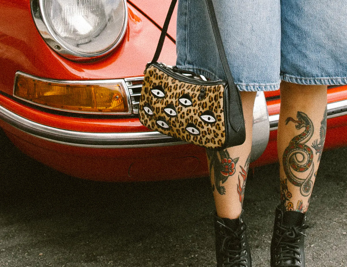 Tattooed woman in denim Bermuda shorts stands by a red vintage car, holding a leopard-print shoulder bag with eye motifs, and wearing black platform combat boots on asphalt.