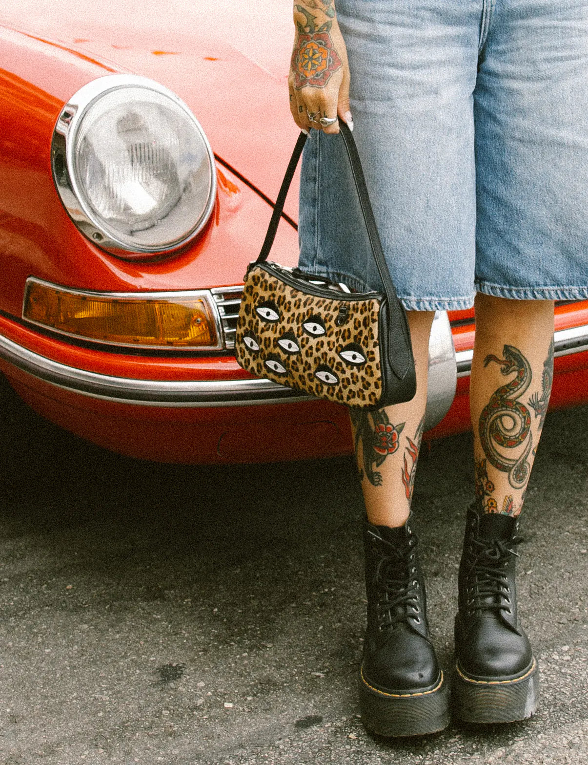 Tattooed woman in denim Bermuda shorts stands by a red vintage car, holding a leopard-print shoulder bag with eye motifs, and wearing black platform combat boots on asphalt.