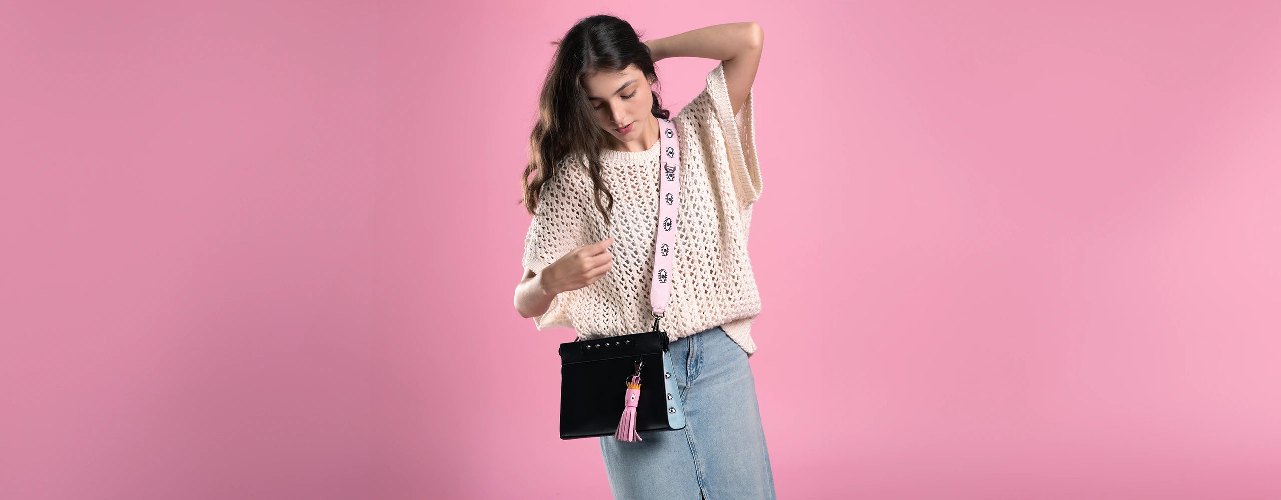 A woman with long brown hair wears a cream-colored knit sweater and a light blue denim skirt, holding a black "Vali Crossbody-handbag" by Min & Mon, a NYC-based brand. The handbag features a pink strap with eye designs and a pink tassel charm accessory.