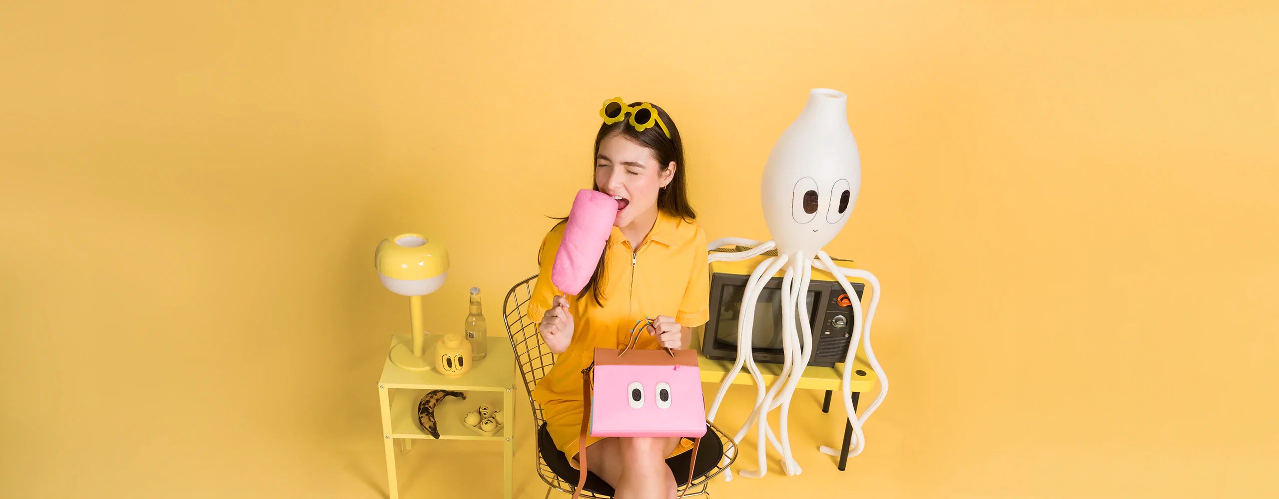 A young woman dressed in a yellow outfit sits on a wire chair in a playful, color-coordinated yellow room. She is biting into a large piece of pink cotton candy while holding a pink handbag with cartoon eyes. She wears sheer socks with small designs and black platform shoes. Behind her are quirky, whimsical props including a white octopus-like lamp with cartoon eyes, a vintage television, a banana, and a bottle of soda on a yellow side table. The overall scene has a surreal, fun, and pop-art aesthetic.