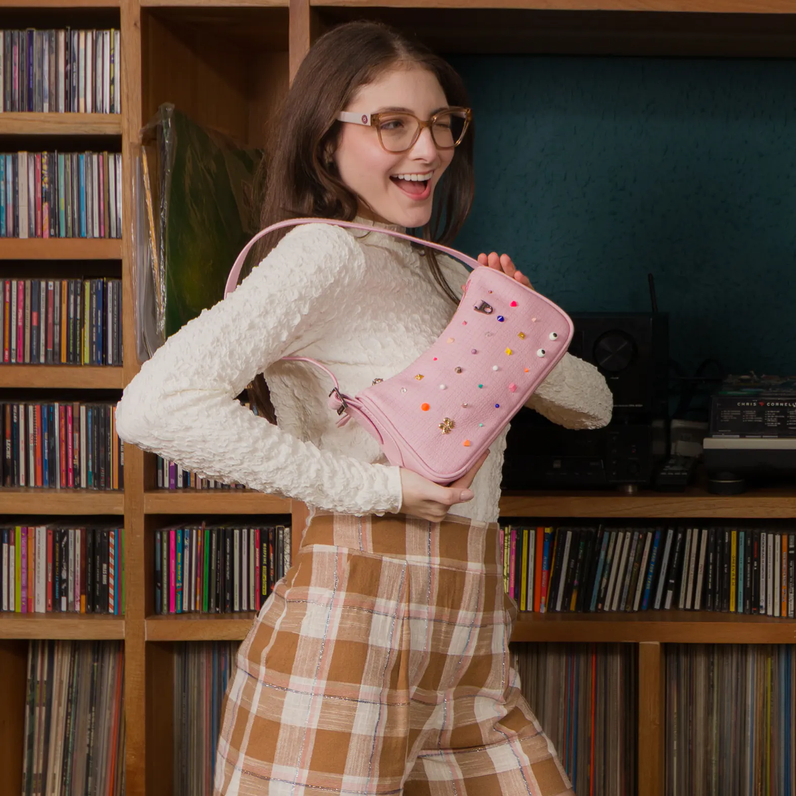 Smiling woman in glasses poses indoors holding a pink shoulder bag adorned with colorful studs and eye motifs, wearing a white textured top and brown plaid shorts.