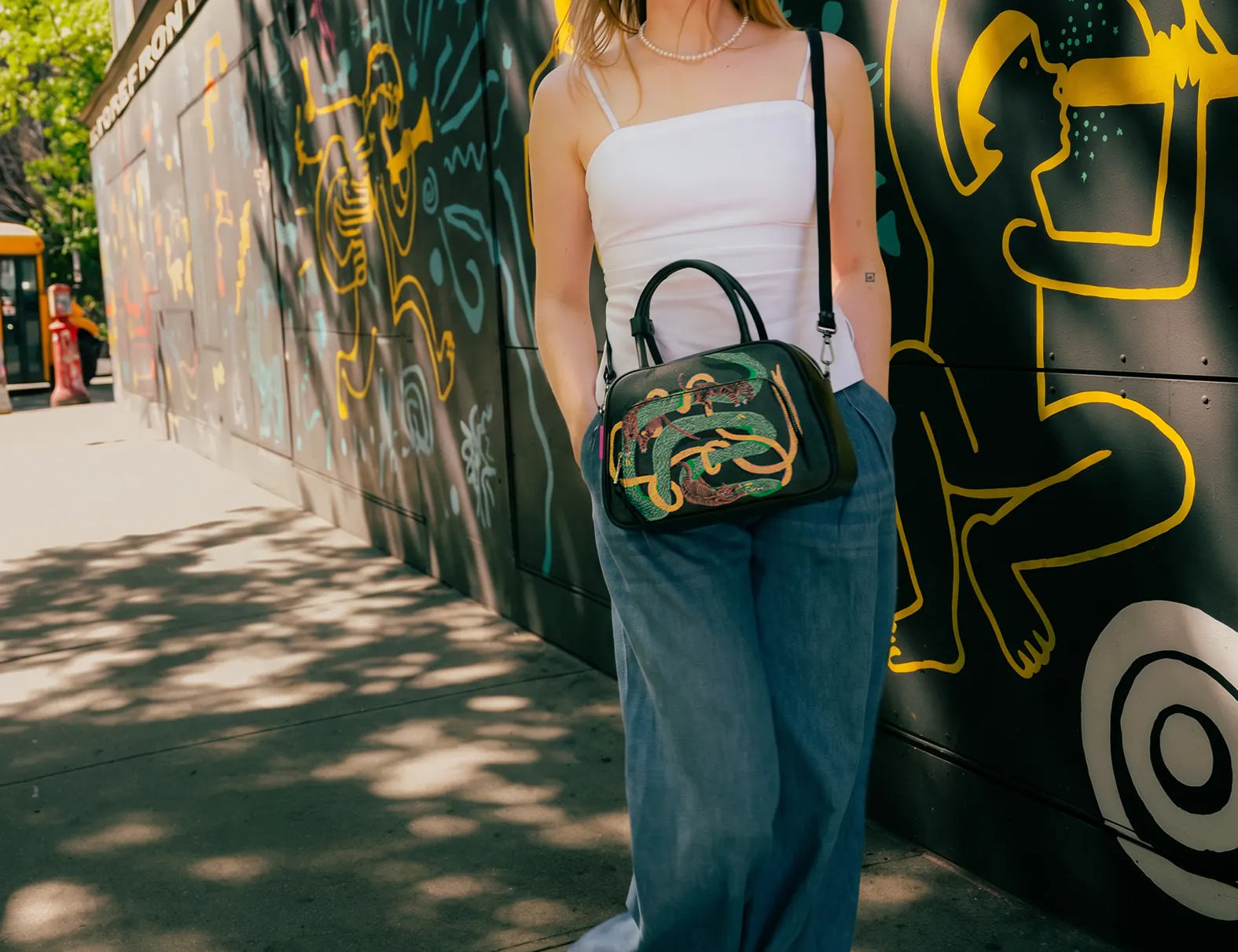 Woman in a white camisole and wide-leg jeans walks along a graffiti-covered wall, wearing sunglasses and carrying a black handbag with colorful embroidered snake design.