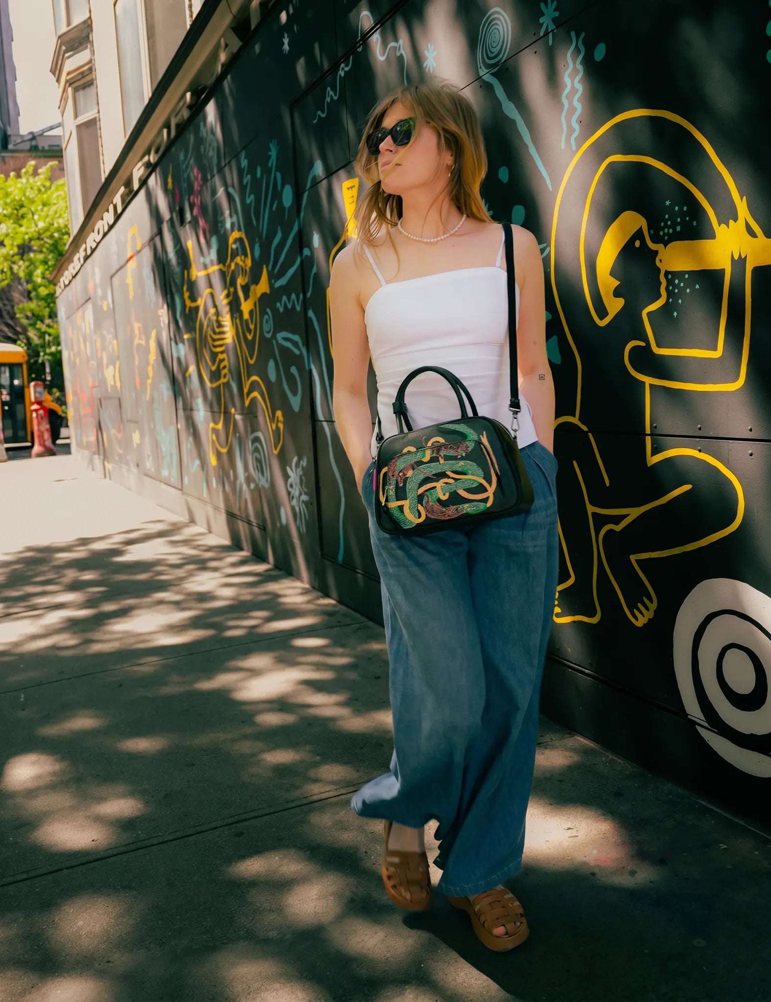 Woman in a white camisole and wide-leg jeans walks along a graffiti-covered wall, wearing sunglasses and carrying a black handbag with colorful embroidered snake design.