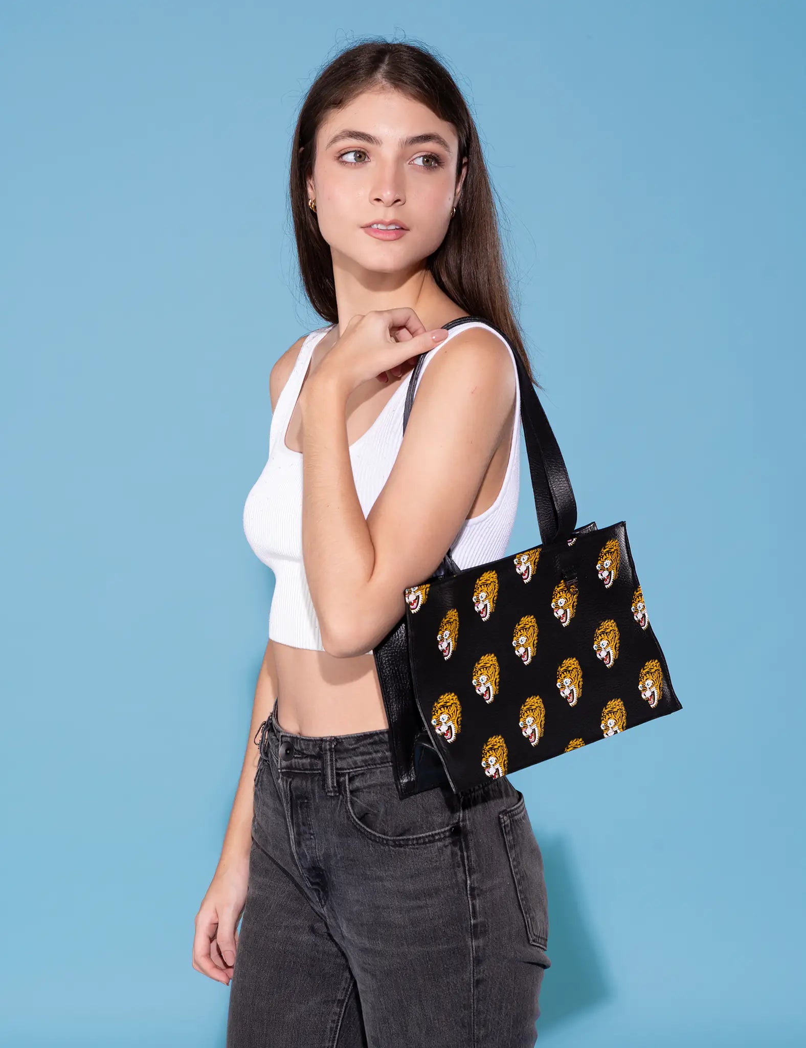 A young woman wearing a white crop top and black jeans is showcasing a Marko tote bag with a black background and multiple colorful tiger head designs. The background is a solid light blue, creating a vibrant contrast.