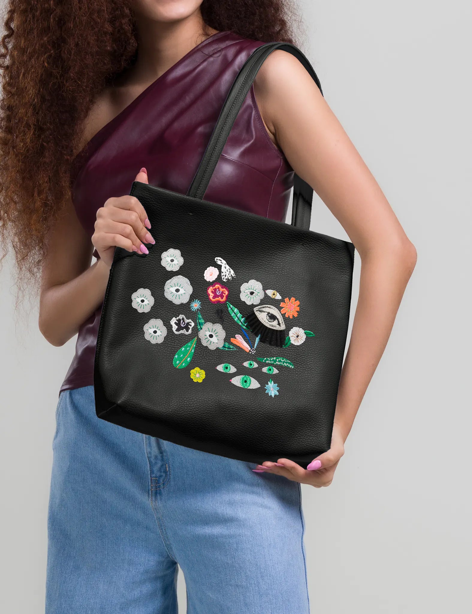 Close-up of a black leather tote bag with colorful embroidered floral and eye motifs, worn over the shoulder by a woman in a burgundy top and light blue jeans.