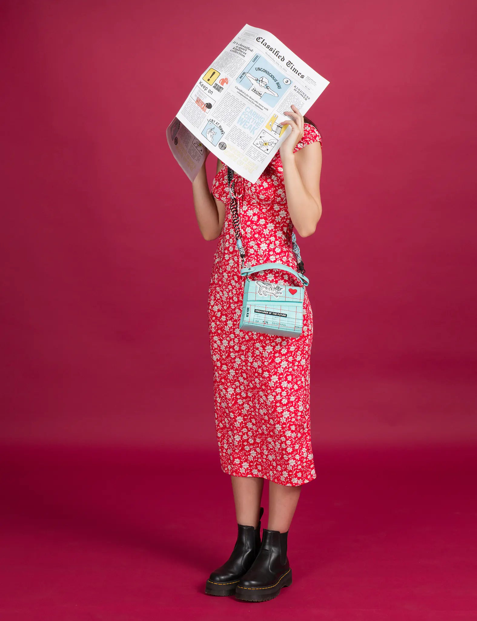 A woman in a red floral dress and black boots stands against a pink backdrop, holding up a newspaper while wearing a turquoise Min & Mon crossbody bag with quirky illustrations and bold lettering.