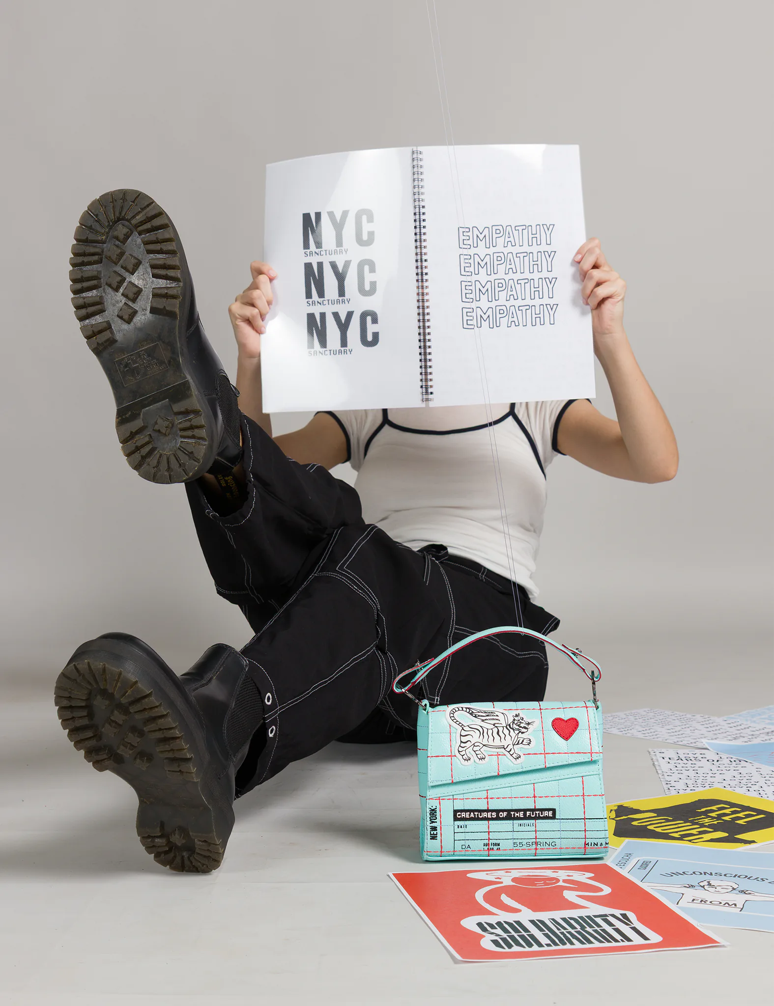 A person in black boots and pants lounges on the floor reading a bold NYC and empathy-themed book, with a turquoise Min & Mon handbag featuring playful graphics and red accents beside them.