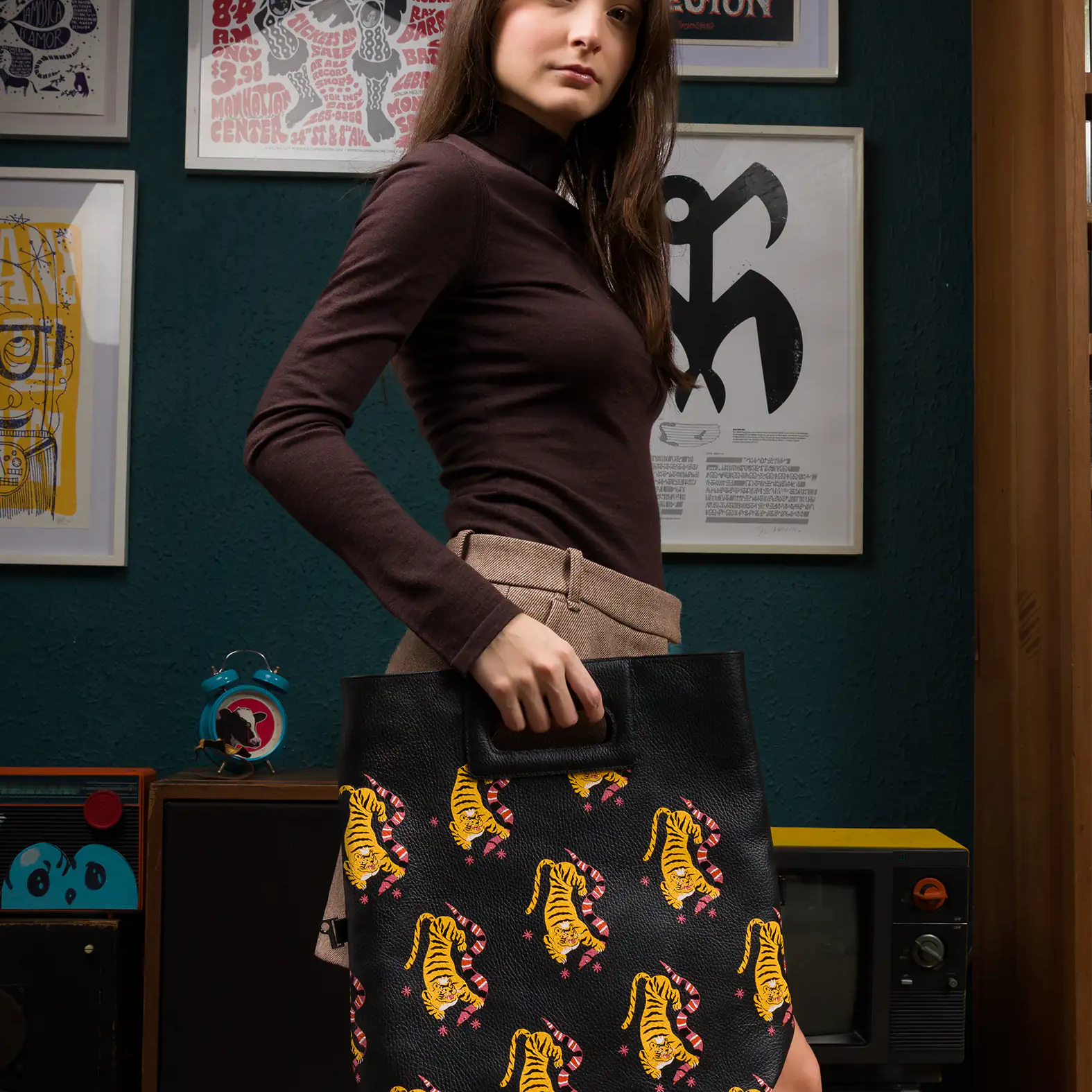 A woman stands indoors holding a black leather tote bag with an printed pattern of yellow tigers. She’s wearing a brown long-sleeve turtleneck top and a tan skirt, in a cozy setting with framed posters and vintage audio equipment behind her.
