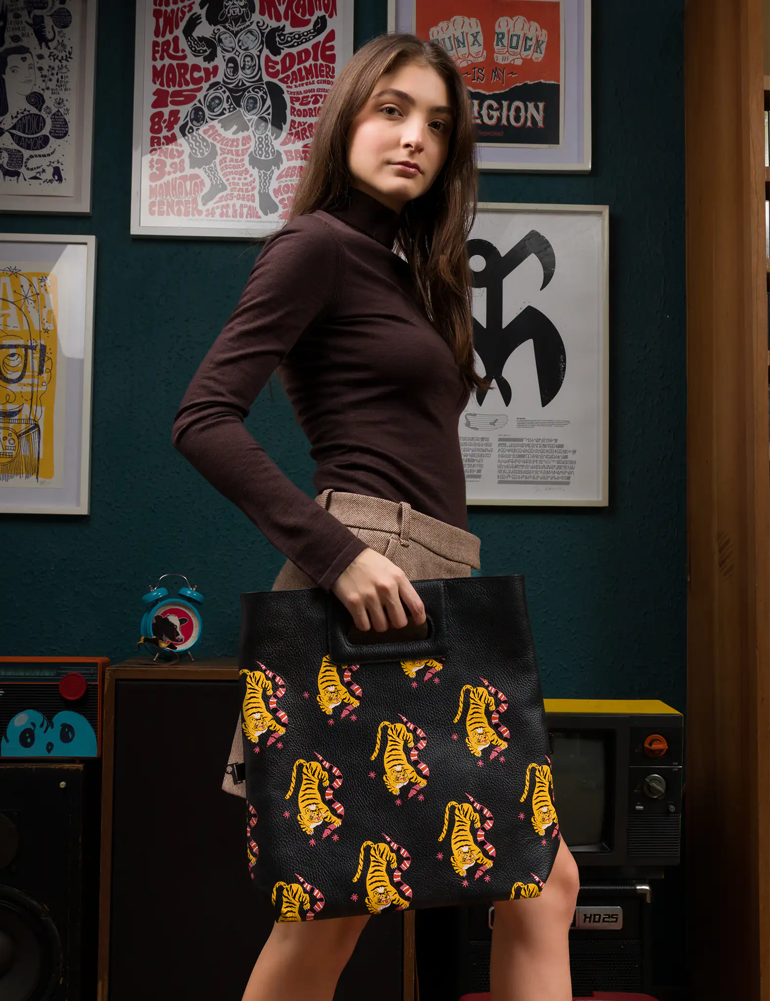 A woman stands indoors holding a black leather tote bag with an printed pattern of yellow tigers. She’s wearing a brown long-sleeve turtleneck top and a tan skirt, in a cozy setting with framed posters and vintage audio equipment behind her.