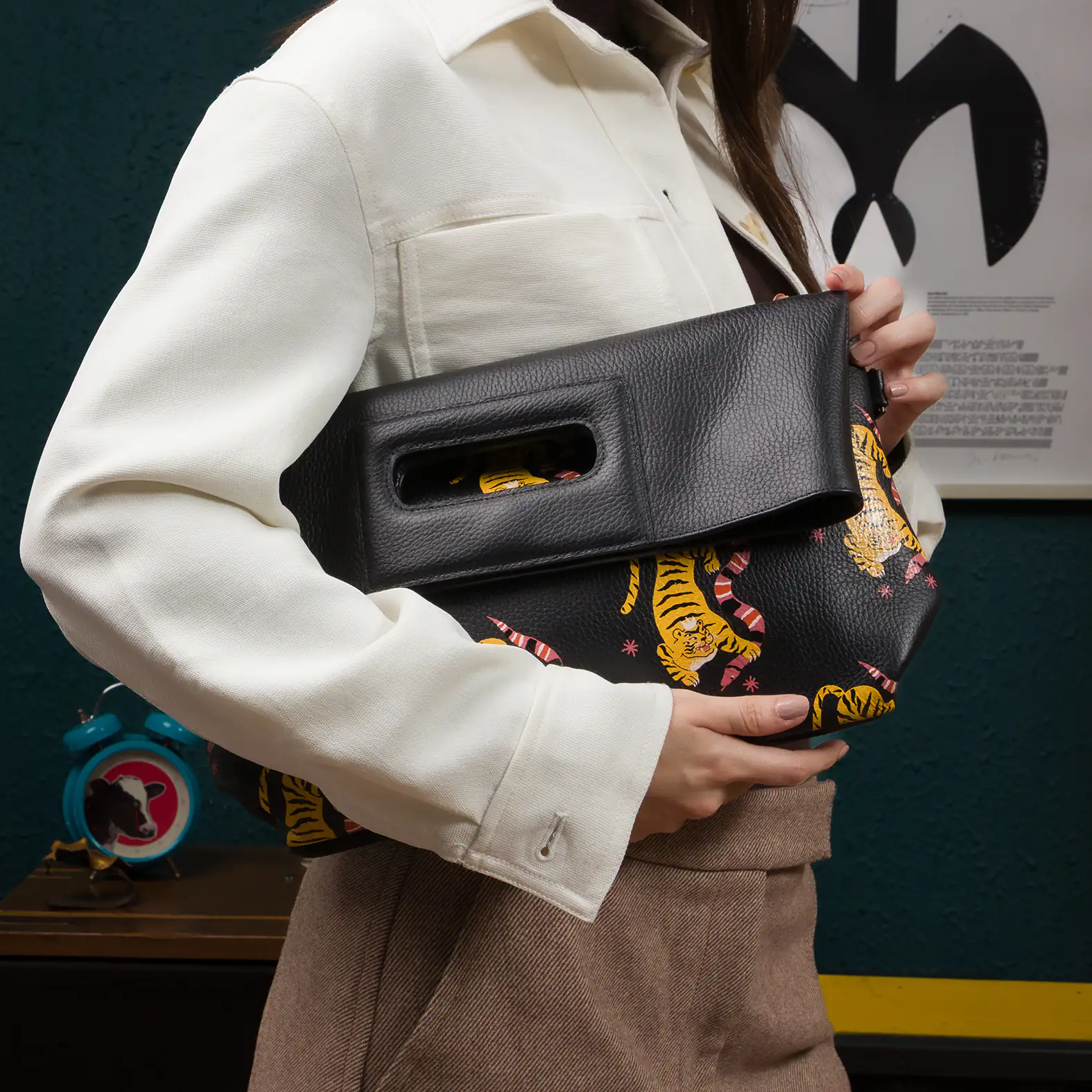 Close-up of a woman holding a black fold-over leather clutch featuring an printed yellow tiger design. She’s wearing a cream button-up jacket, brown skirt, and dark turtleneck in a retro-inspired room.