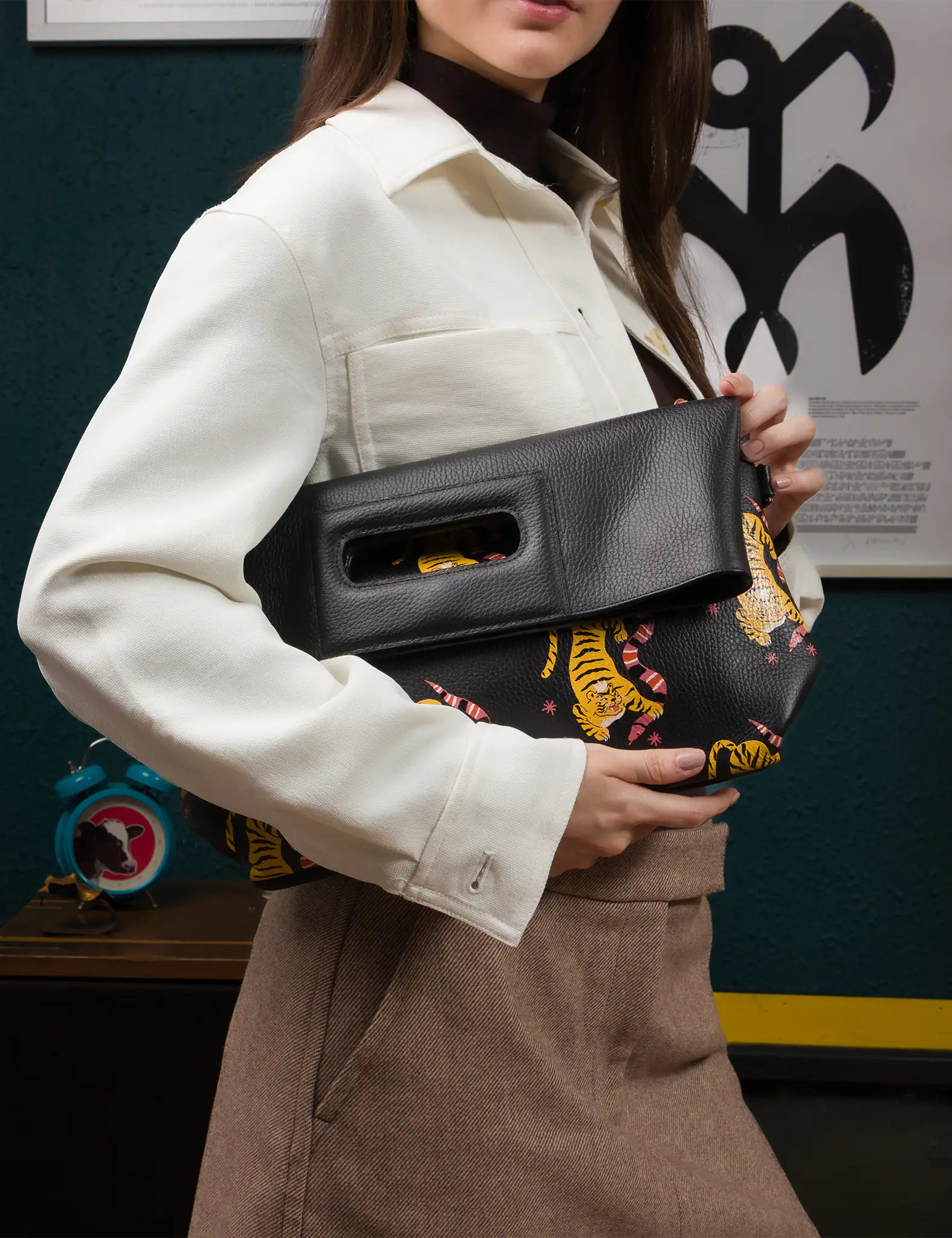 Close-up of a woman holding a black fold-over leather clutch featuring an printed yellow tiger design. She’s wearing a cream button-up jacket, brown skirt, and dark turtleneck in a retro-inspired room.