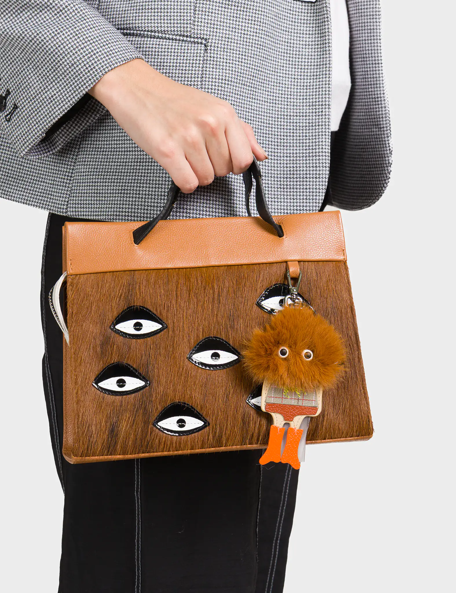 Close-up of a person holding a brown and tan handbag with black-and-white eye motifs and a fluffy brown keychain charm shaped like a quirky character.