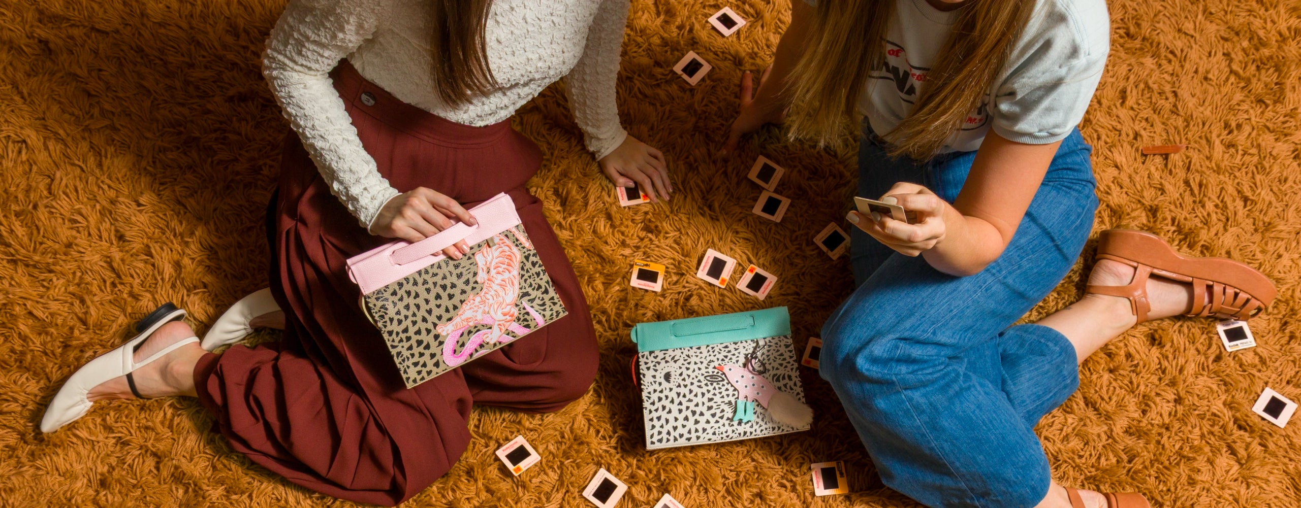Two women sitting on a retro shag rug with Min & Mon animal print handbags from the Retrotiger Collection, surrounded by vintage film slides.