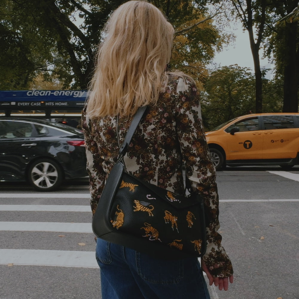 A blonde woman in a patterned blouse and blue jeans walks along a tree-lined New York City street, showcasing a black crossbody bag with colorful designs near a food cart.