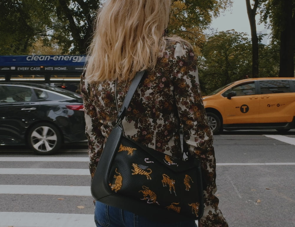 A blonde woman in a patterned blouse and blue jeans walks along a tree-lined New York City street, showcasing a black crossbody bag with colorful designs near a food cart.