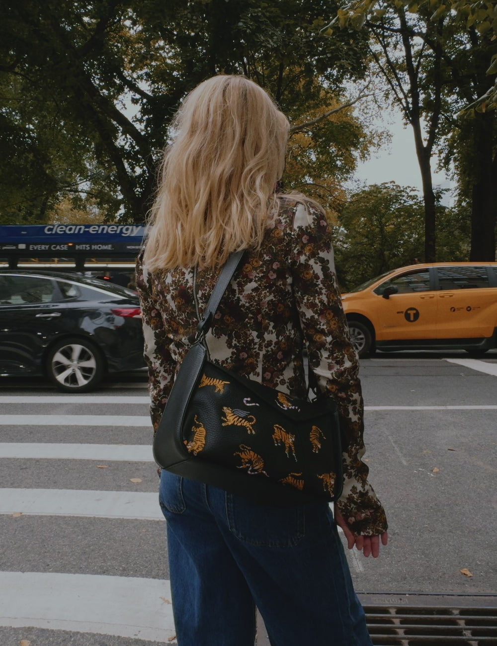 A blonde woman in a patterned blouse and blue jeans walks along a tree-lined New York City street, showcasing a black crossbody bag with colorful designs near a food cart.