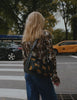 A blonde woman in a patterned blouse and blue jeans walks along a tree-lined New York City street, showcasing a black crossbody bag with colorful designs near a food cart.