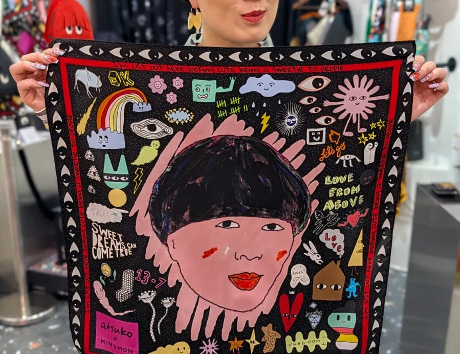A woman in a retail store holds up a square black scarf featuring a colorful whimsical illustration of a face with a black wig, surrounded by doodles, stars, and text.