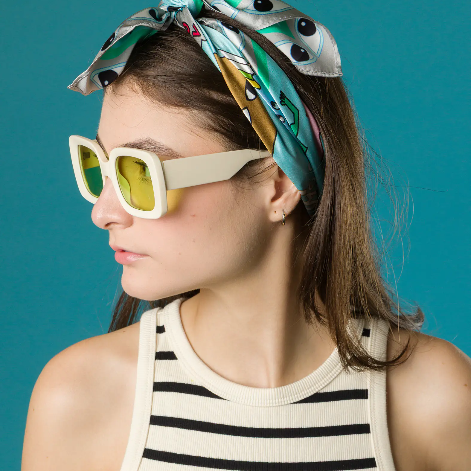 A woman wears a turquoise Min & Mon silk scarf with eye motifs tied as a headband, paired with bold yellow-lensed sunglasses and a striped tank top against a teal background.