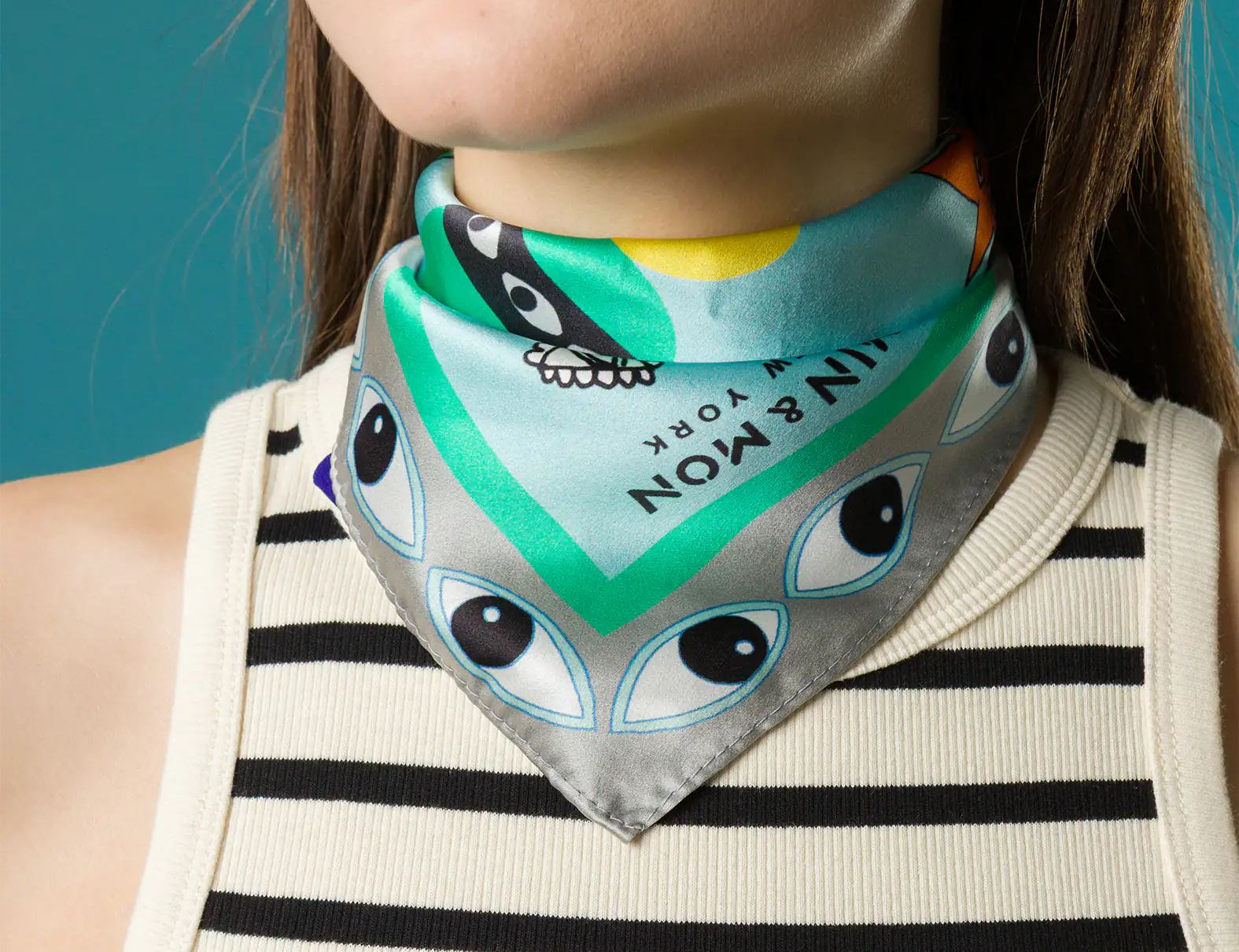 Close-up of a woman wearing a Min & Mon silk scarf tied around her neck, featuring bold turquoise, silver, and black eye motifs and the brand’s logo, styled with a striped tank top.
