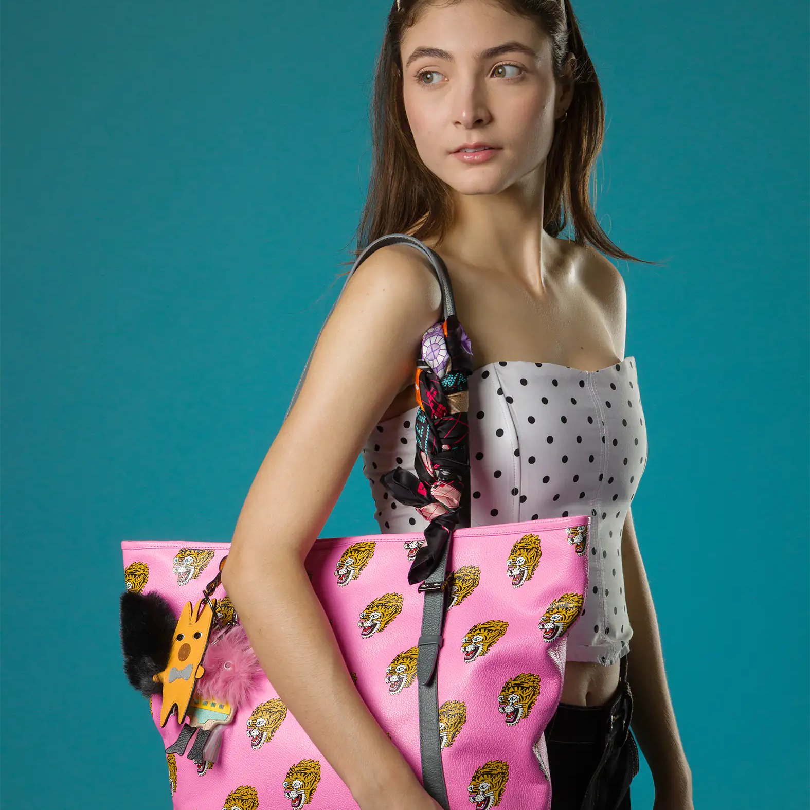 A woman in a white polka-dot corset top carries a bold pink Min & Mon tote bag decorated with repeating tiger faces and playful charms, styled with a printed scarf on the strap.
