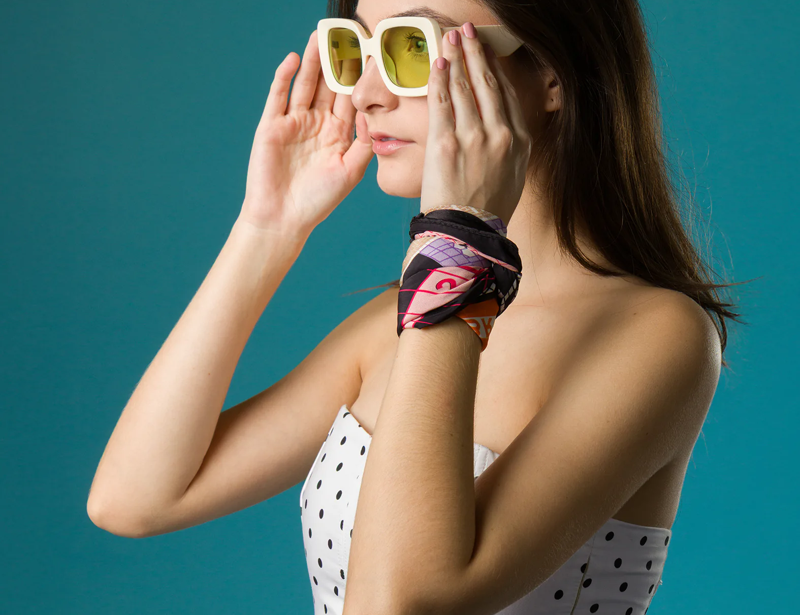 A woman in a white polka-dot corset top adjusts her bold yellow-lensed sunglasses, wearing a colorful Min & Mon silk scarf wrapped stylishly around her wrist as an accessory.