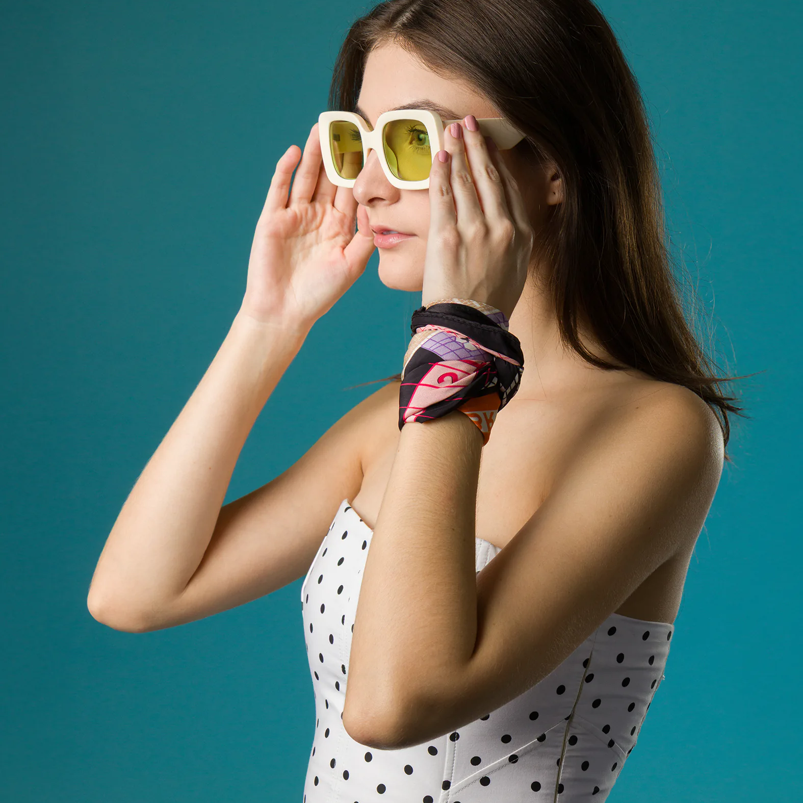 A woman in a white polka-dot corset top adjusts her bold yellow-lensed sunglasses, wearing a colorful Min & Mon silk scarf wrapped stylishly around her wrist as an accessory.