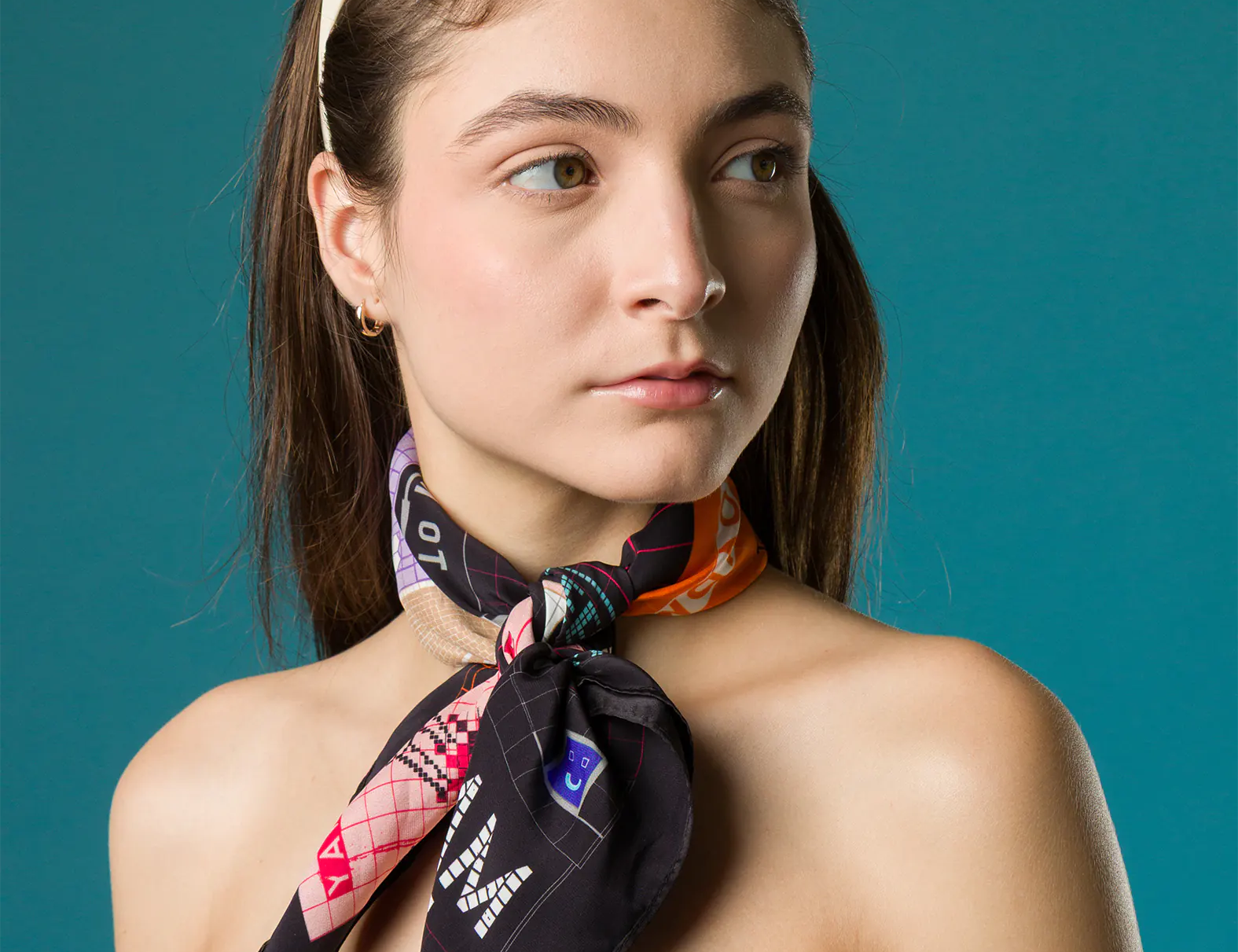 A woman wears a colorful Min & Mon silk scarf tied around her neck, paired with a white polka-dot top and sunglasses resting on her head, set against a teal background.