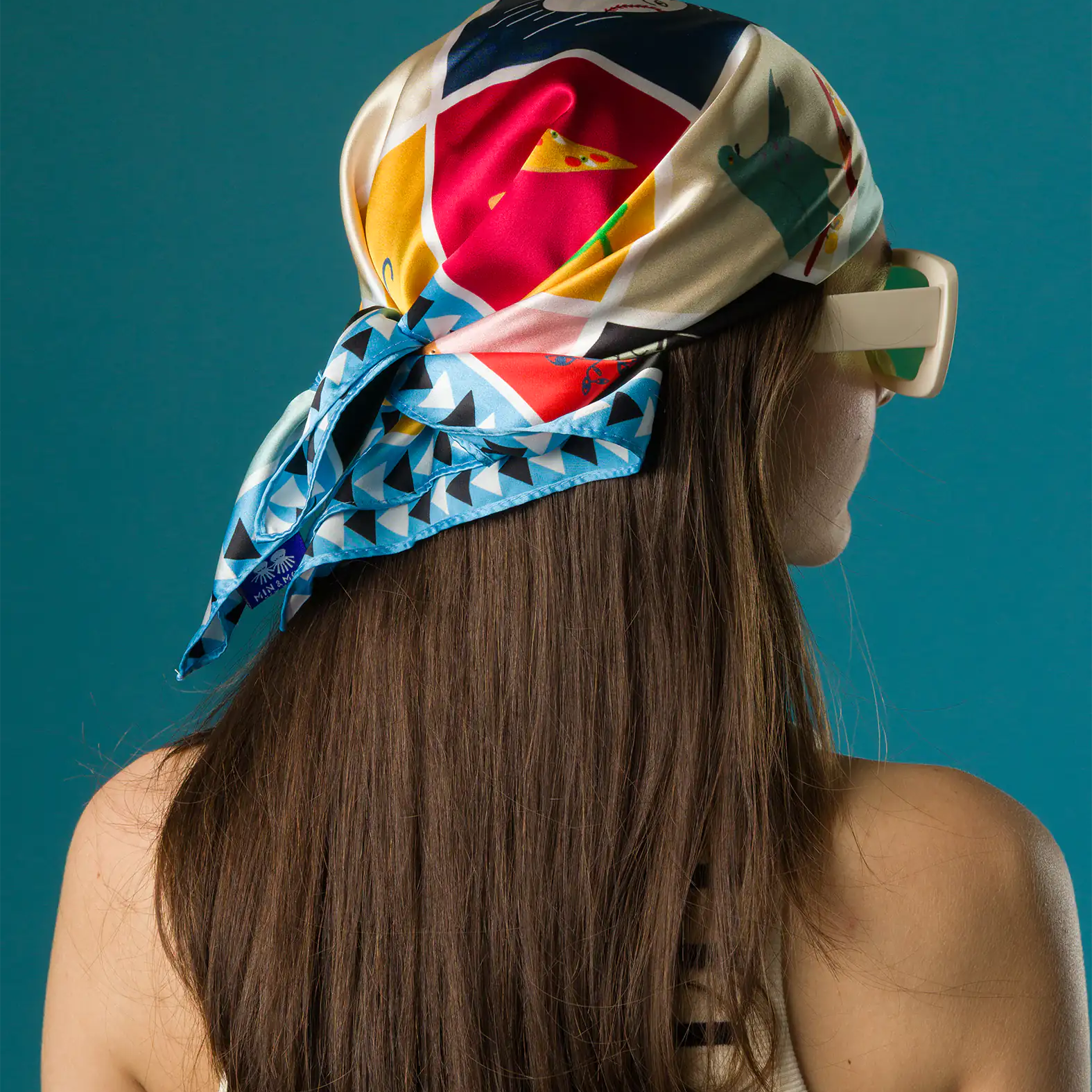 A woman wears a colorful Min & Mon silk scarf as a head wrap, featuring playful illustrations and blue geometric trim, styled with a striped tank top and bold white sunglasses.