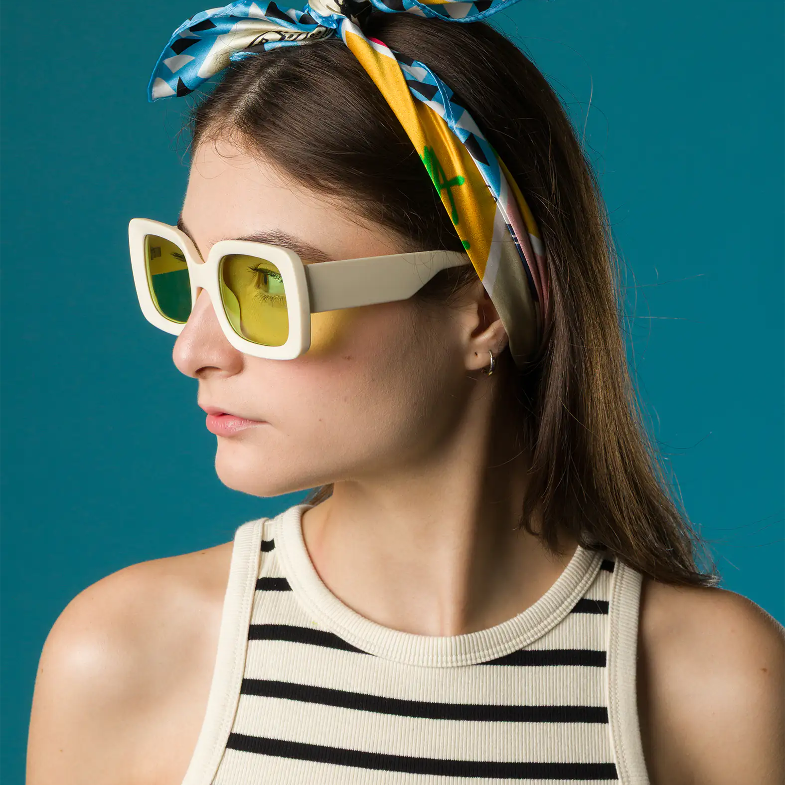 A woman in a striped tank top wears a colorful silk Min & Mon scarf tied as a headband with a bow, paired with oversized yellow-lensed sunglasses against a teal background.