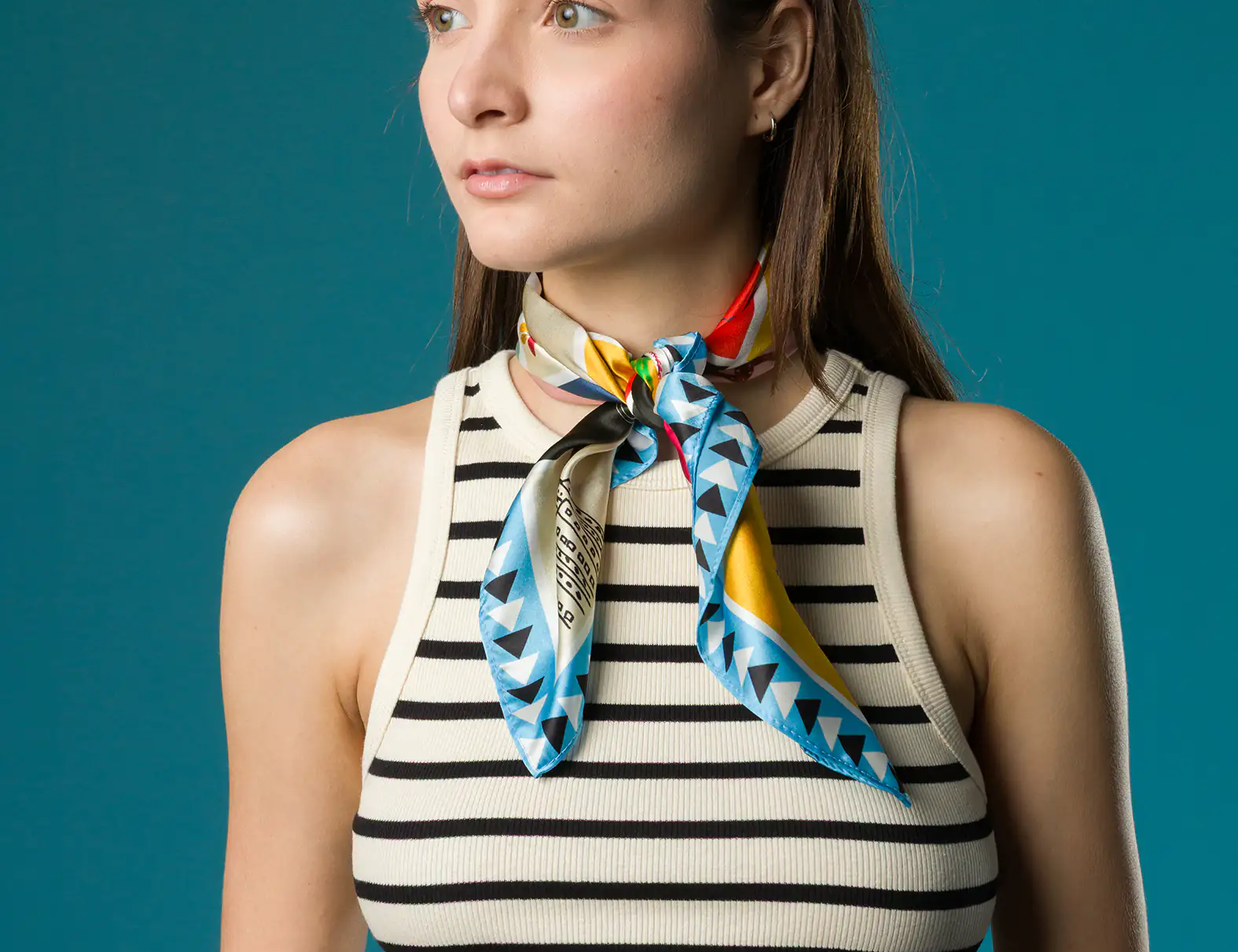A woman in a striped crop top wears a vibrant Min & Mon silk scarf tied neatly around her neck, featuring blue triangle patterns and bold red and yellow accents, with sunglasses resting on her head.