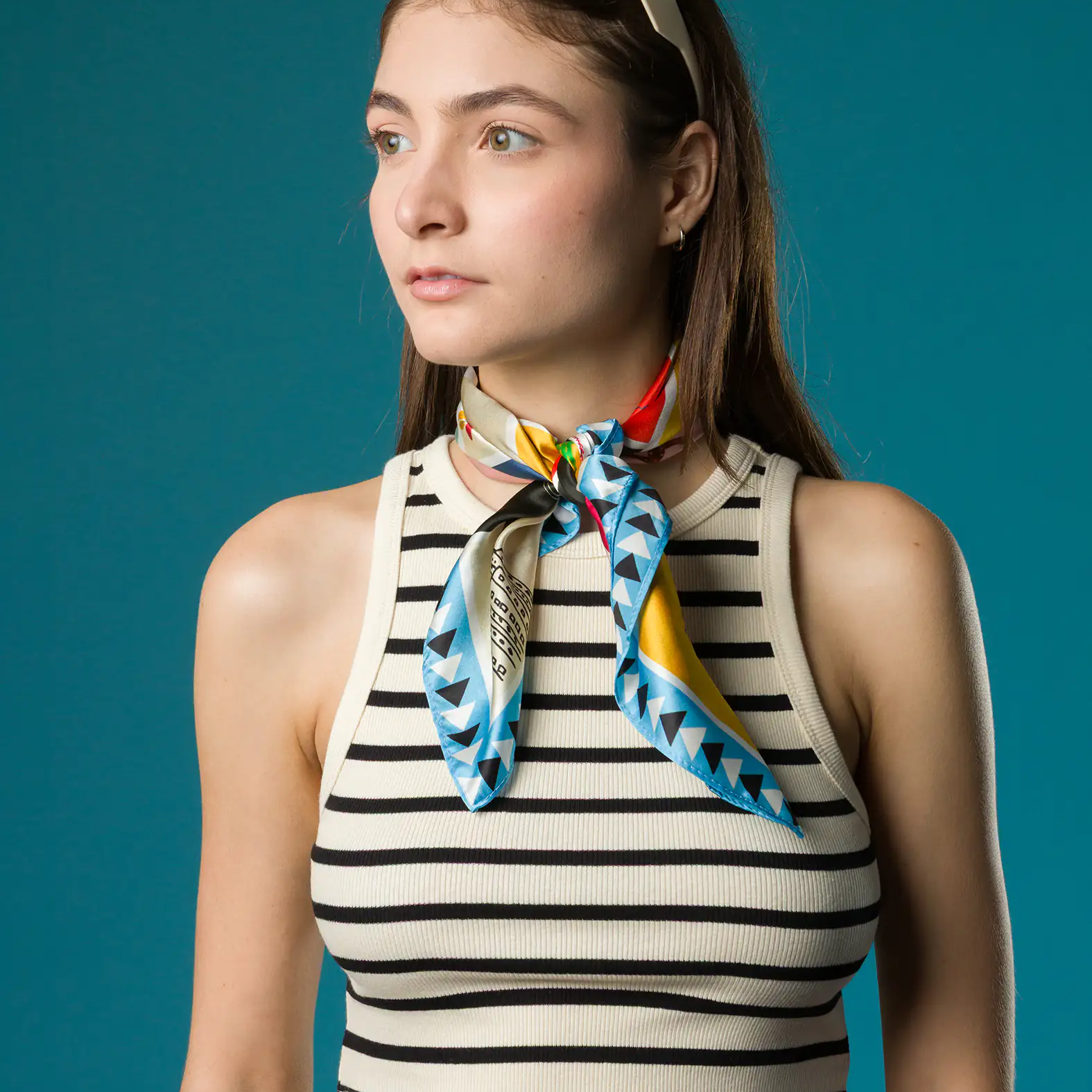 A woman in a striped crop top wears a vibrant Min & Mon silk scarf tied neatly around her neck, featuring blue triangle patterns and bold red and yellow accents, with sunglasses resting on her head.