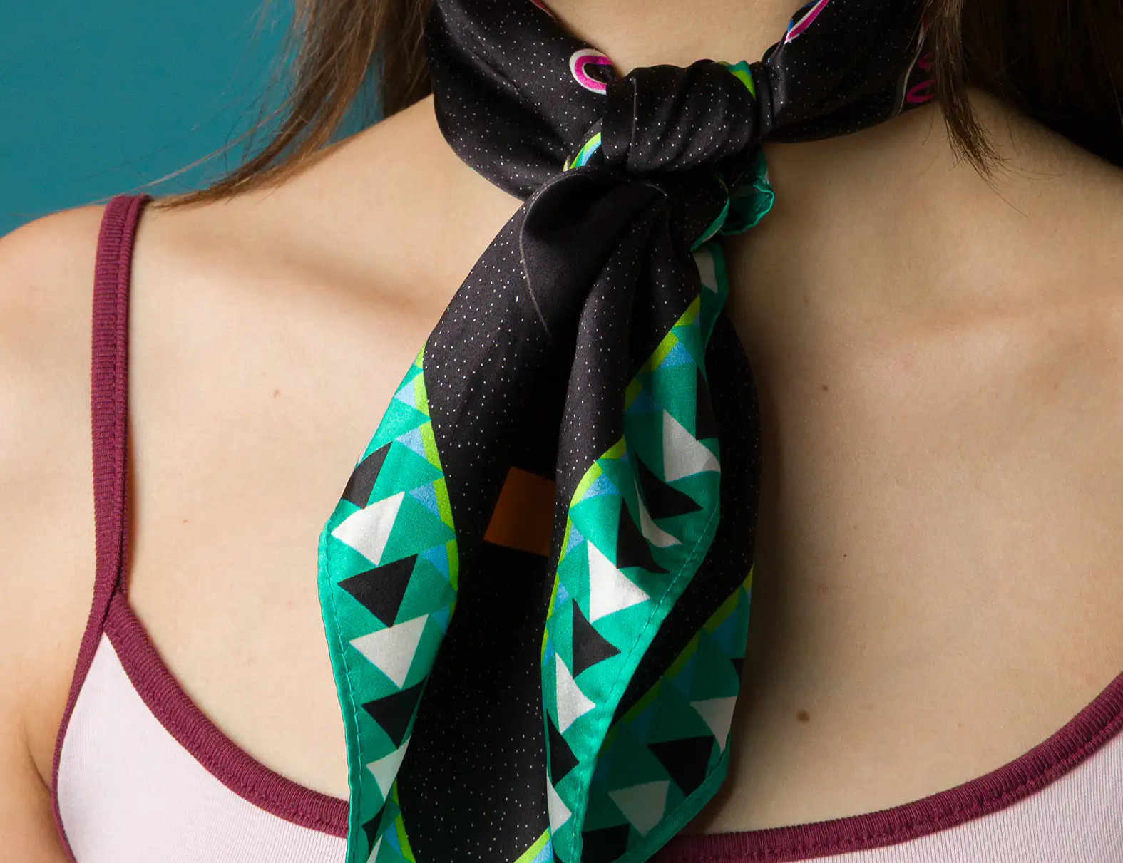 Close-up of a woman wearing a stylish black silk scarf featuring teal, white, and green triangle patterns, tied at the front over a light pink tank top with burgundy trim.