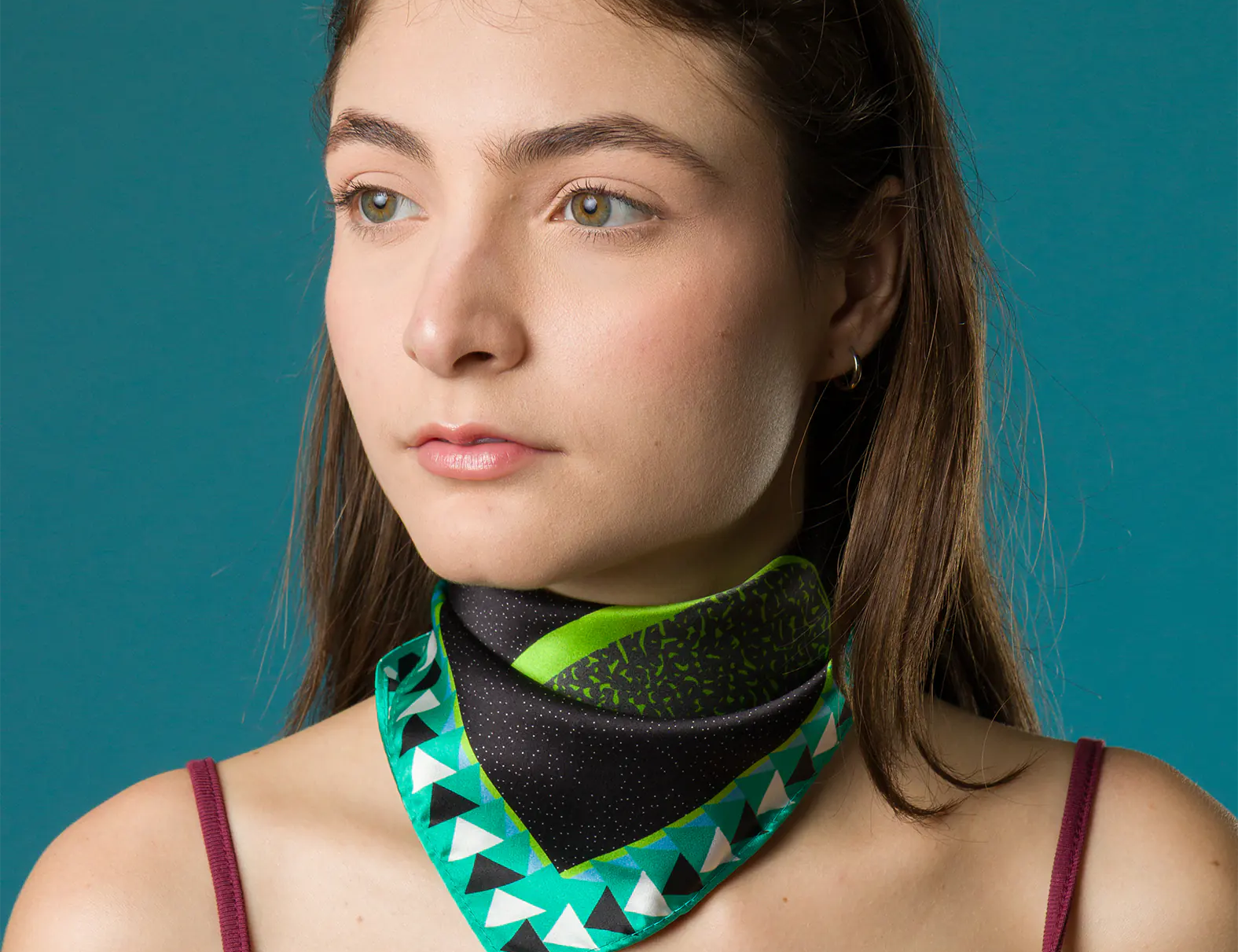 A woman with red sunglasses atop her head wears a silk scarf with vibrant green, teal, and black geometric patterns tied around her neck, paired with a burgundy-trimmed tank top.