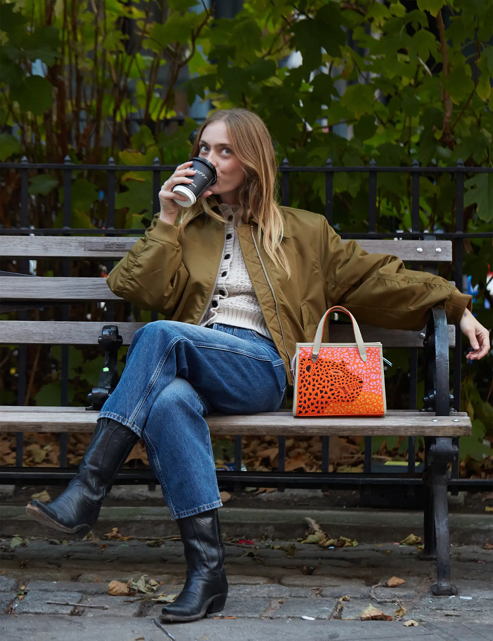 A woman sitting on a park bench sipping coffee, dressed in jeans, a beige sweater, and an olive bomber jacket, with an orange patterned handbag beside her.