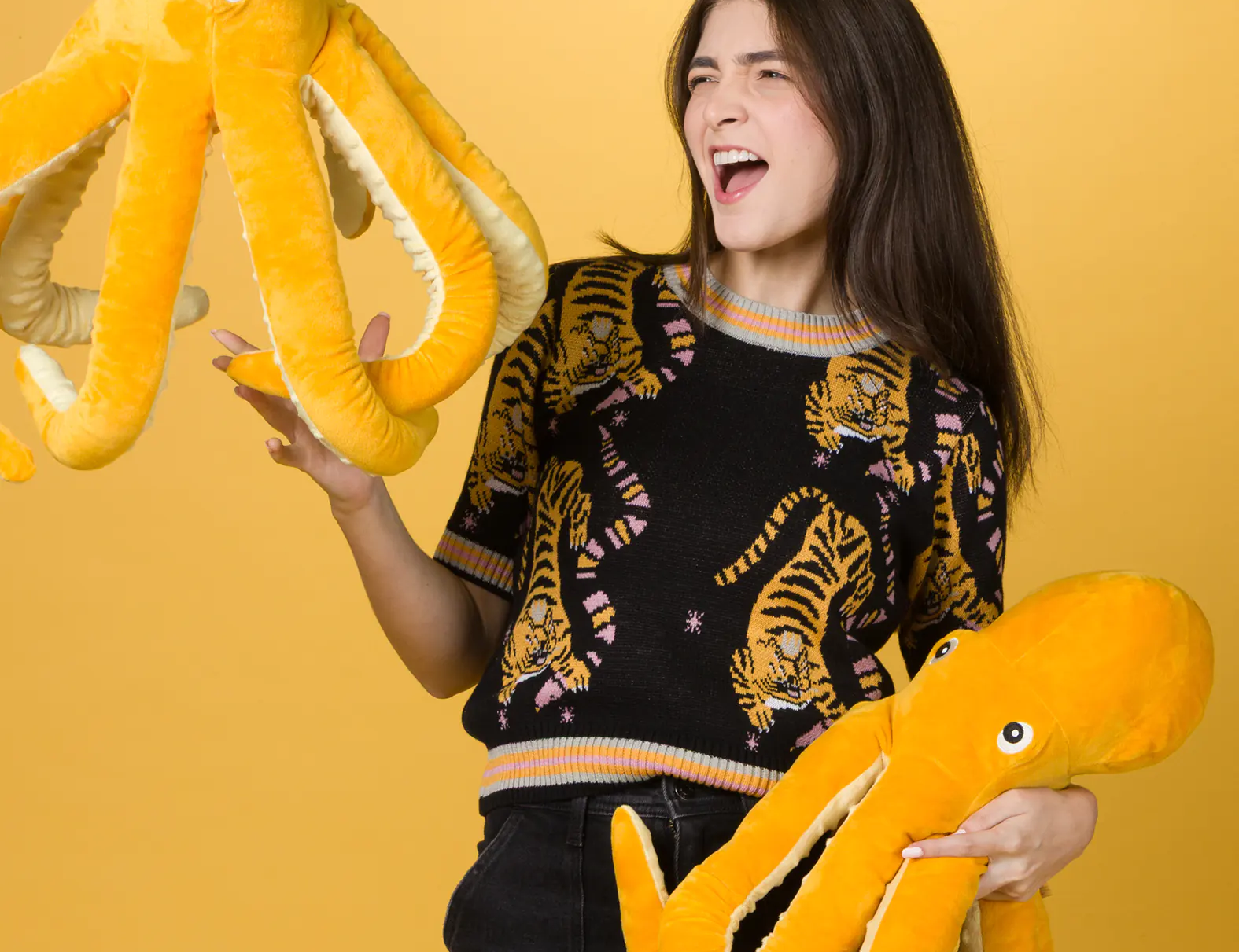 Woman smiling and holding two large yellow octopus plush toys against a solid yellow background, wearing a black sweater with a colorful tiger print and black high-waisted pants.