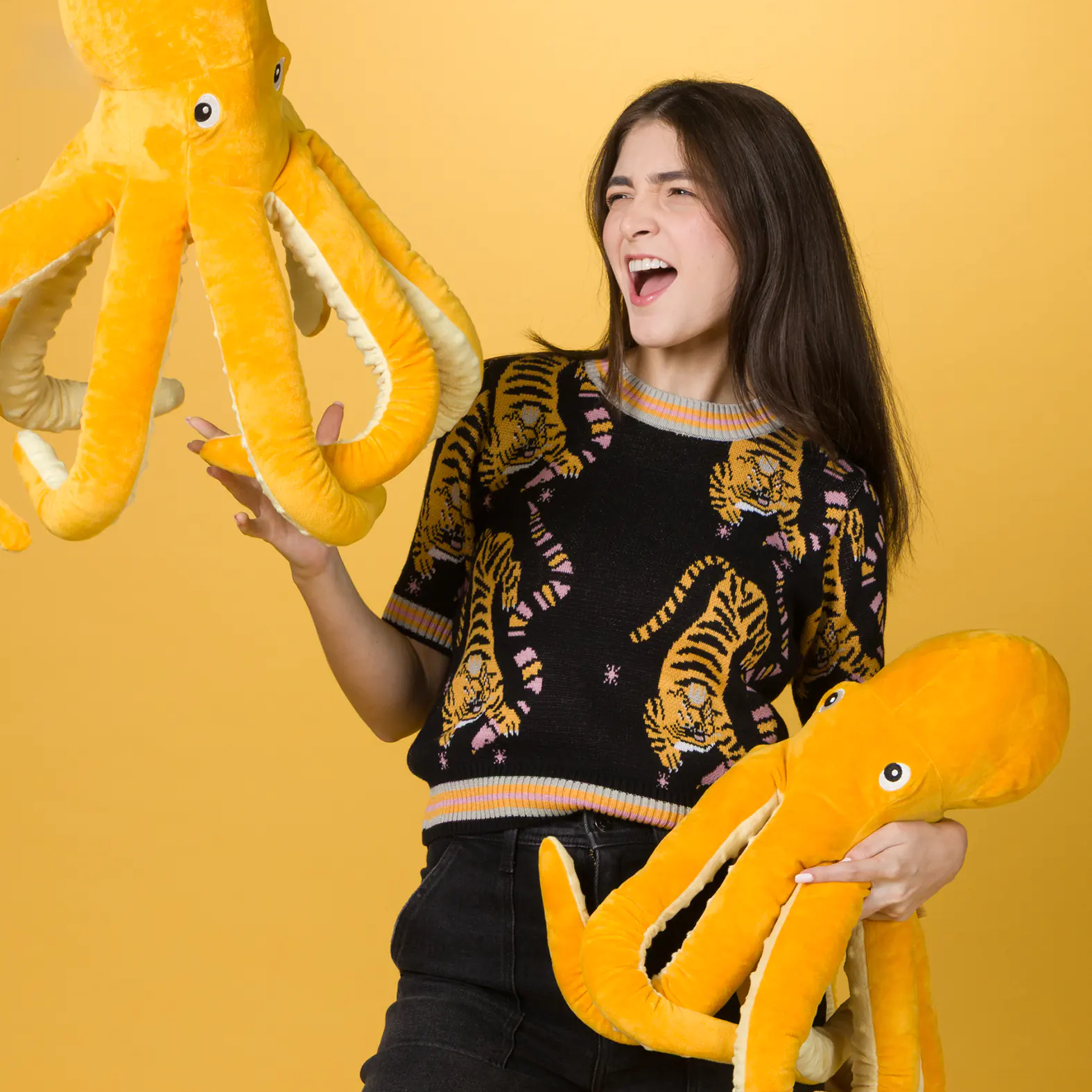 Woman smiling and holding two large yellow octopus plush toys against a solid yellow background, wearing a black sweater with a colorful tiger print and black high-waisted pants.
