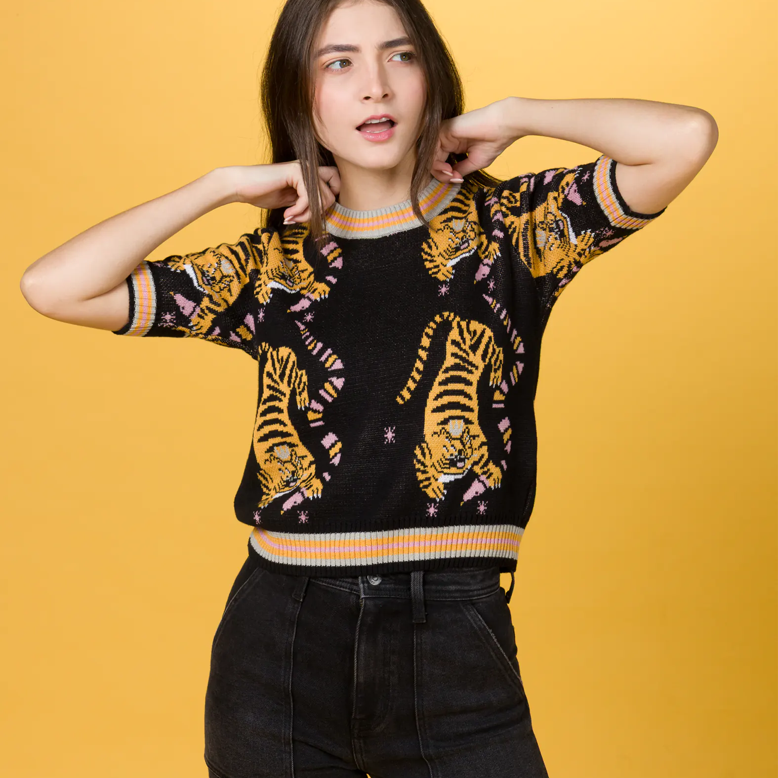 Woman standing against a yellow background, wearing a black short-sleeve sweater with a colorful tiger print and high-waisted black jeans, posing with her hands near her neck and a thoughtful expression.