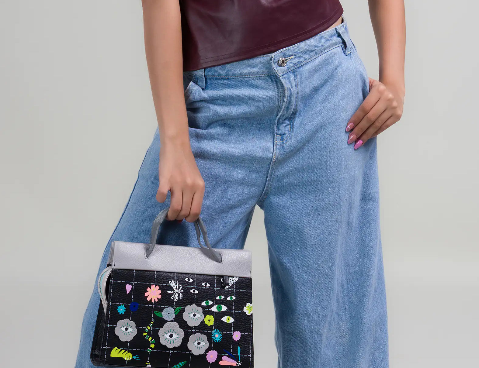 Woman wearing a burgundy sleeveless top and light blue wide-leg jeans holds a structured handbag featuring a black base with colorful floral and eye motifs and a gray top handle.
