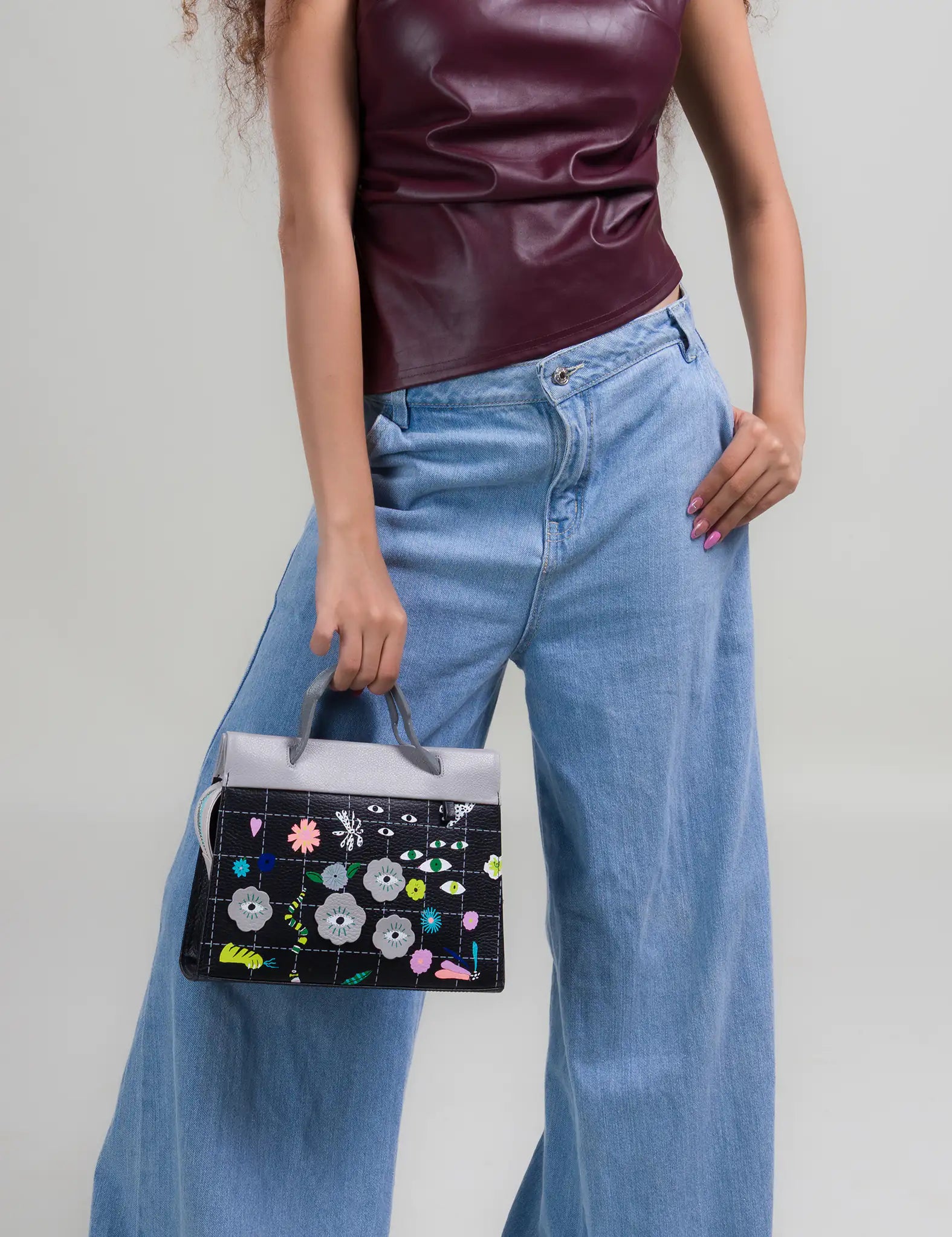 Woman wearing a burgundy sleeveless top and light blue wide-leg jeans holds a structured handbag featuring a black base with colorful floral and eye motifs and a gray top handle.