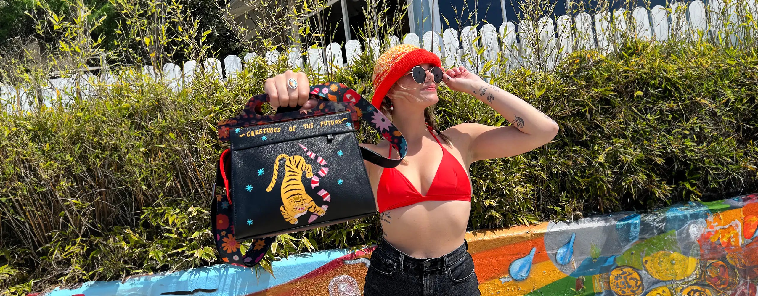 A woman wearing a red bikini top, a red crocheted hat, and sunglasses poses outdoors, holding a "Vali plastic handle crossbody handbag" by Min & Mon, a NYC-based brand. The black handbag features a colorful strap and a vibrant tiger and snake print with the text "Creatures of the Future."