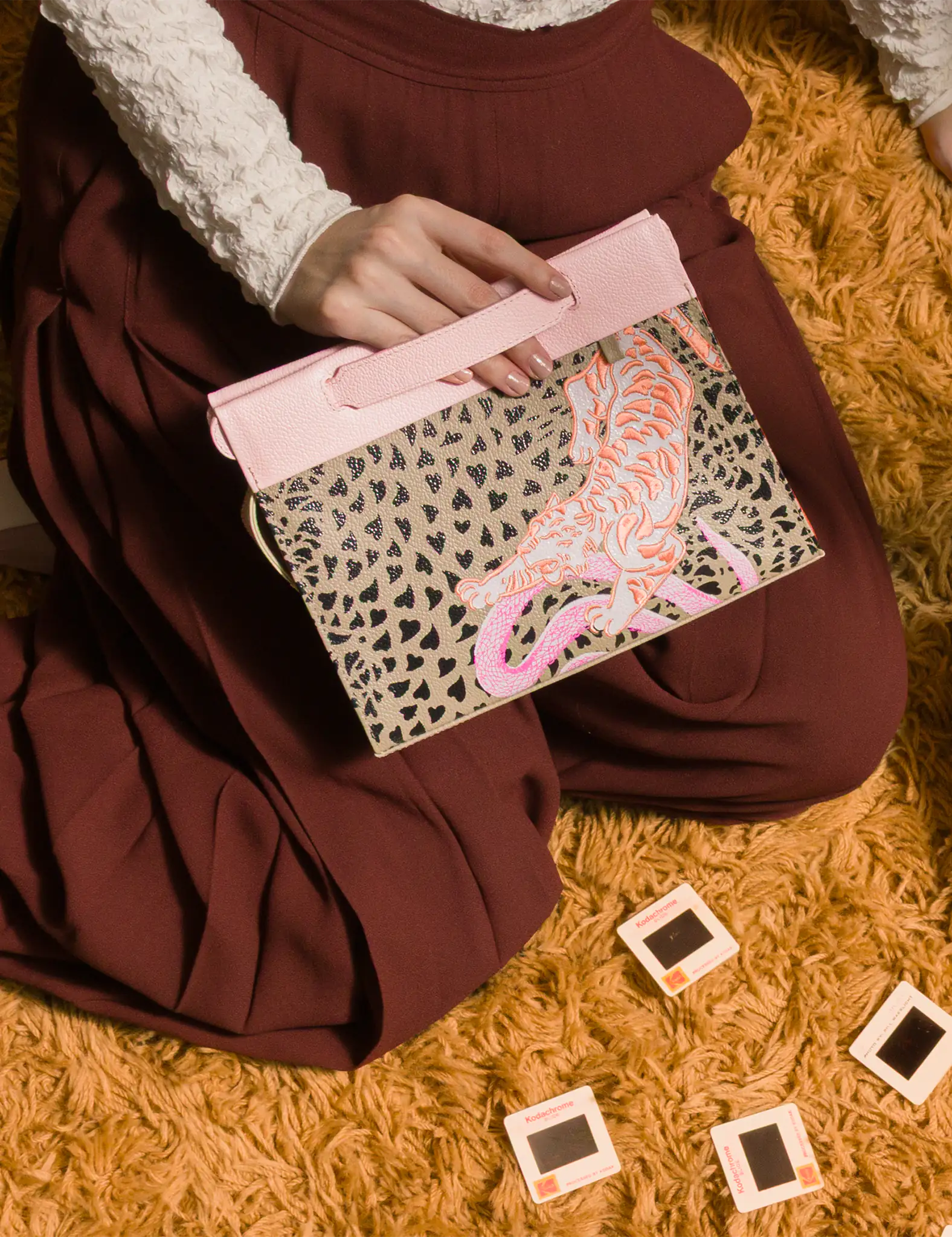 Close-up of a person sitting on a carpeted floor, holding a beige and pink clutch bag with a heart pattern and embroidered pink tiger design.