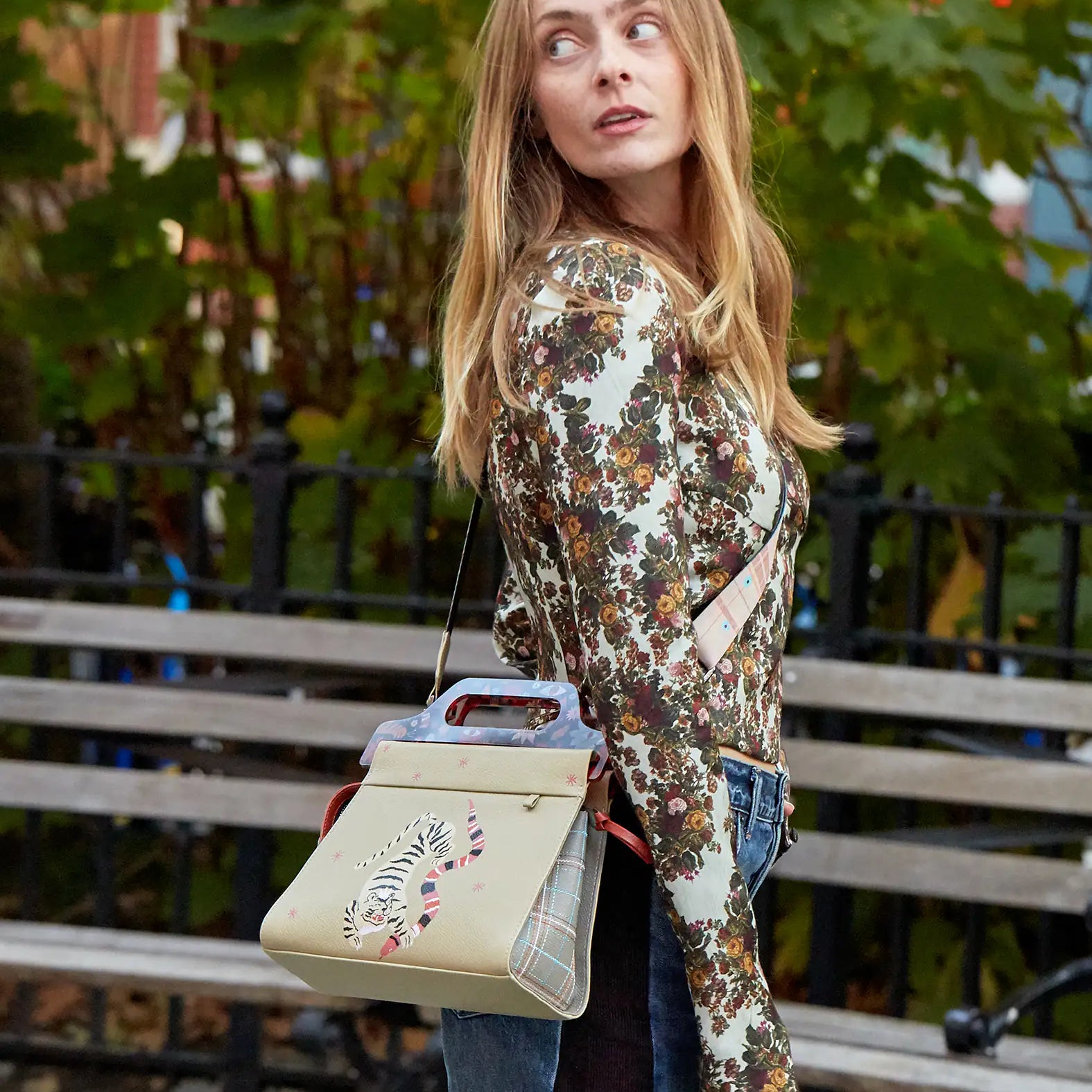 Woman in a floral top and jeans looks over her shoulder in a park, wearing a beige handbag with a tiger design and structured handle, set against trees and a bench.