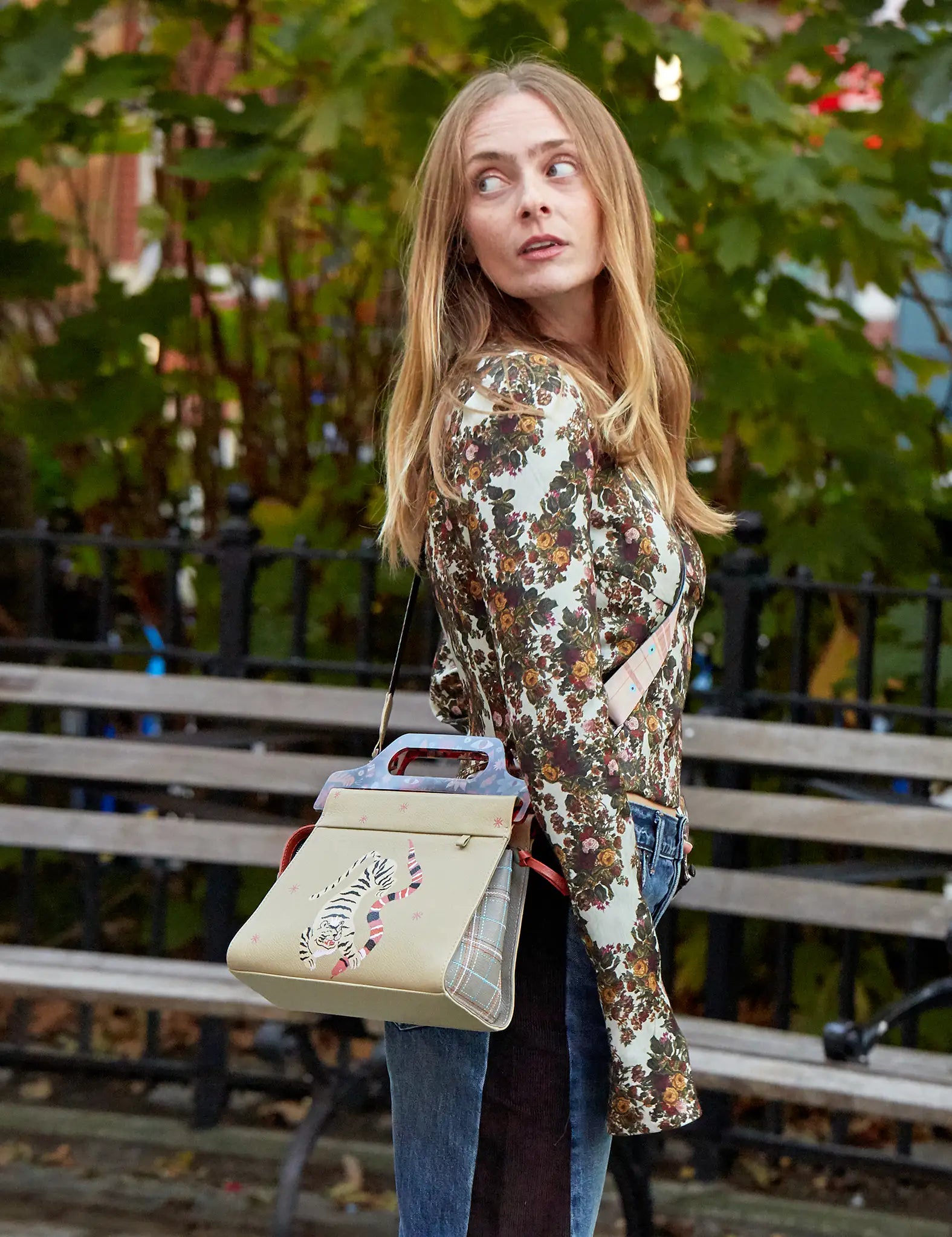 Woman in a floral top and jeans looks over her shoulder in a park, wearing a beige handbag with a tiger design and structured handle, set against trees and a bench.