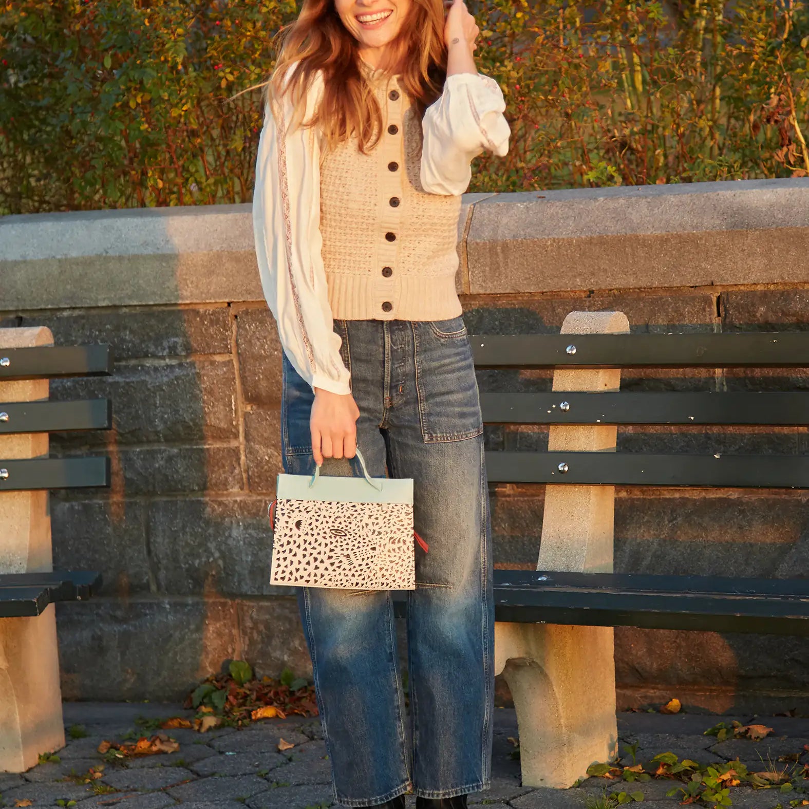 Woman standing outdoors holding a patterned handbag, wearing a beige cardigan and blue jeans.
